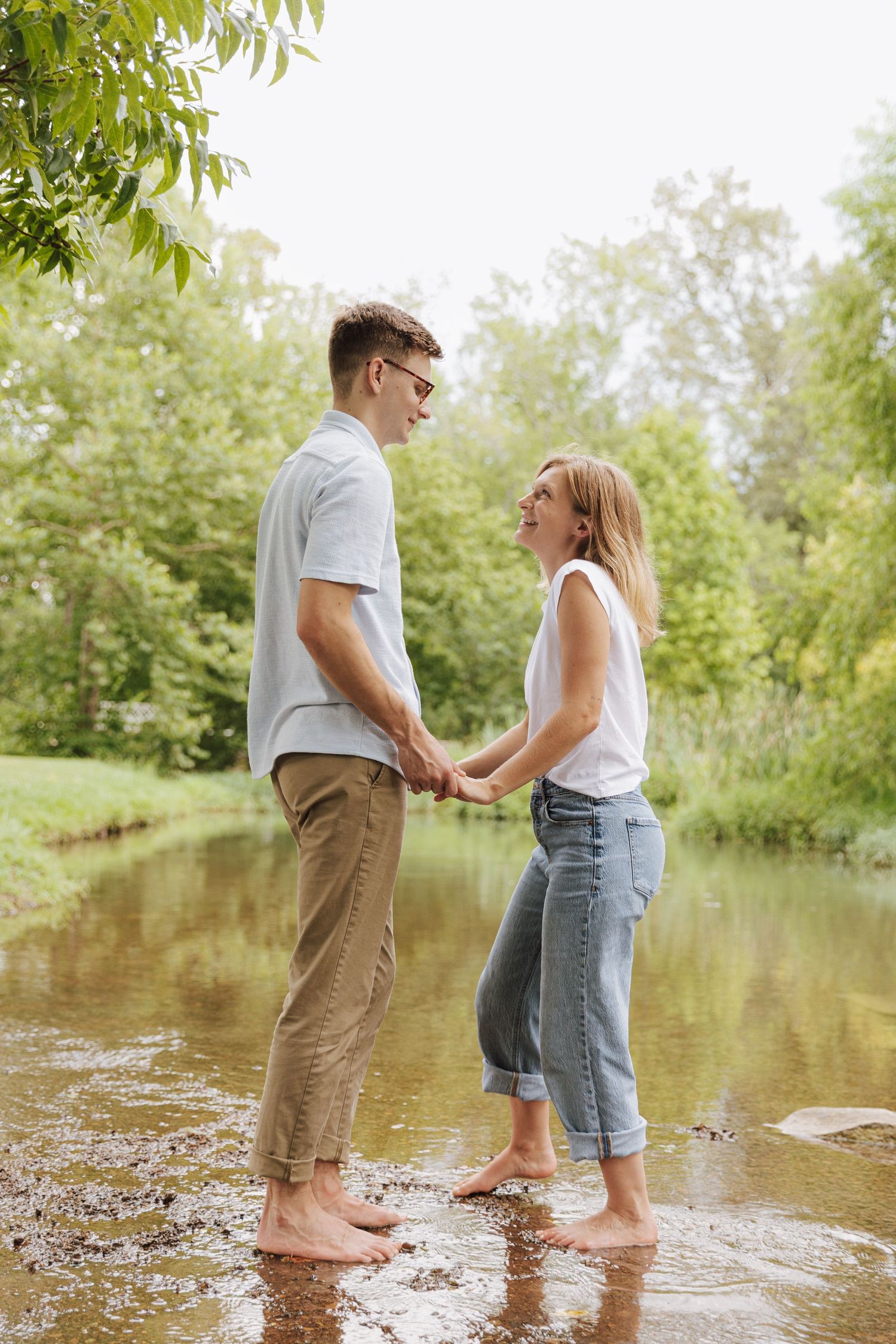 A couple stands barefoot in shallow water holding hands during an outdoor photoshoot.