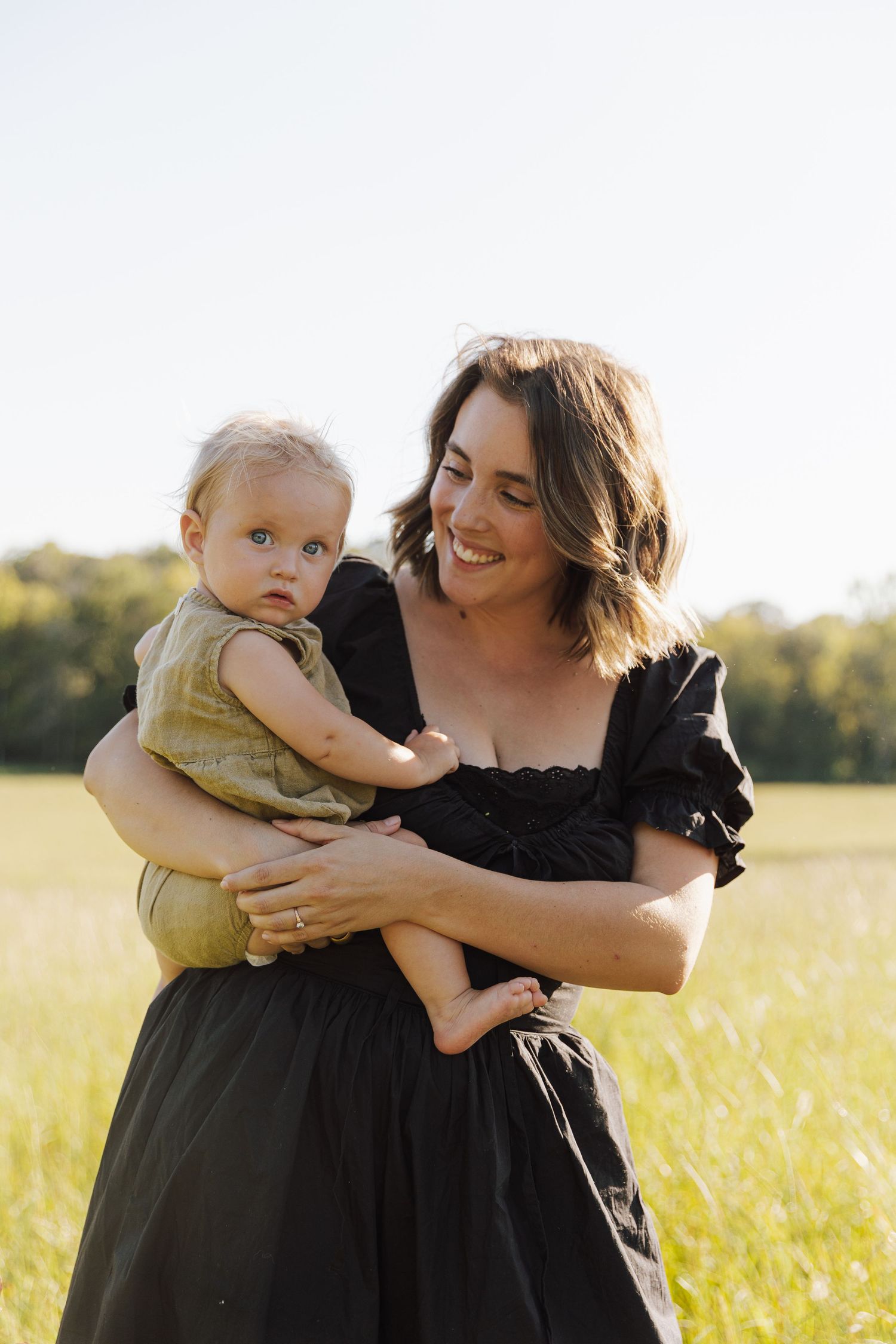 A mom in a black dress holds her baby in a sunlit meadow during golden hour.
