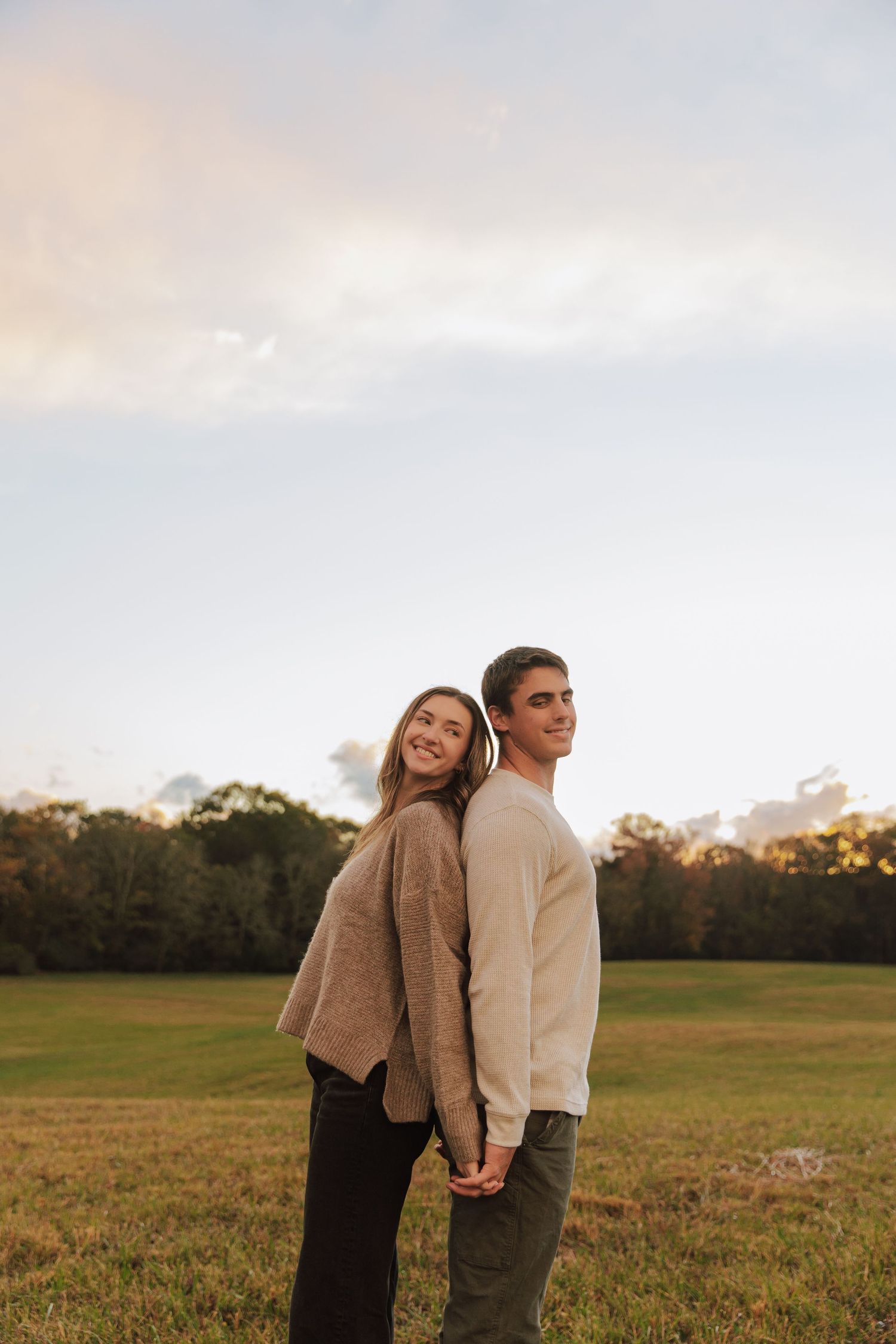 A couple stands back-to-back in a grassy field at sunset wearing cozy sweaters.