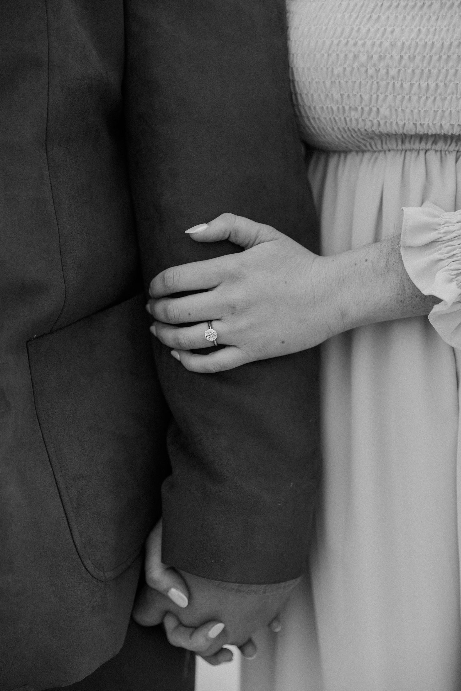A close-up black and white photo of hands with engagement ring against formal attire.
