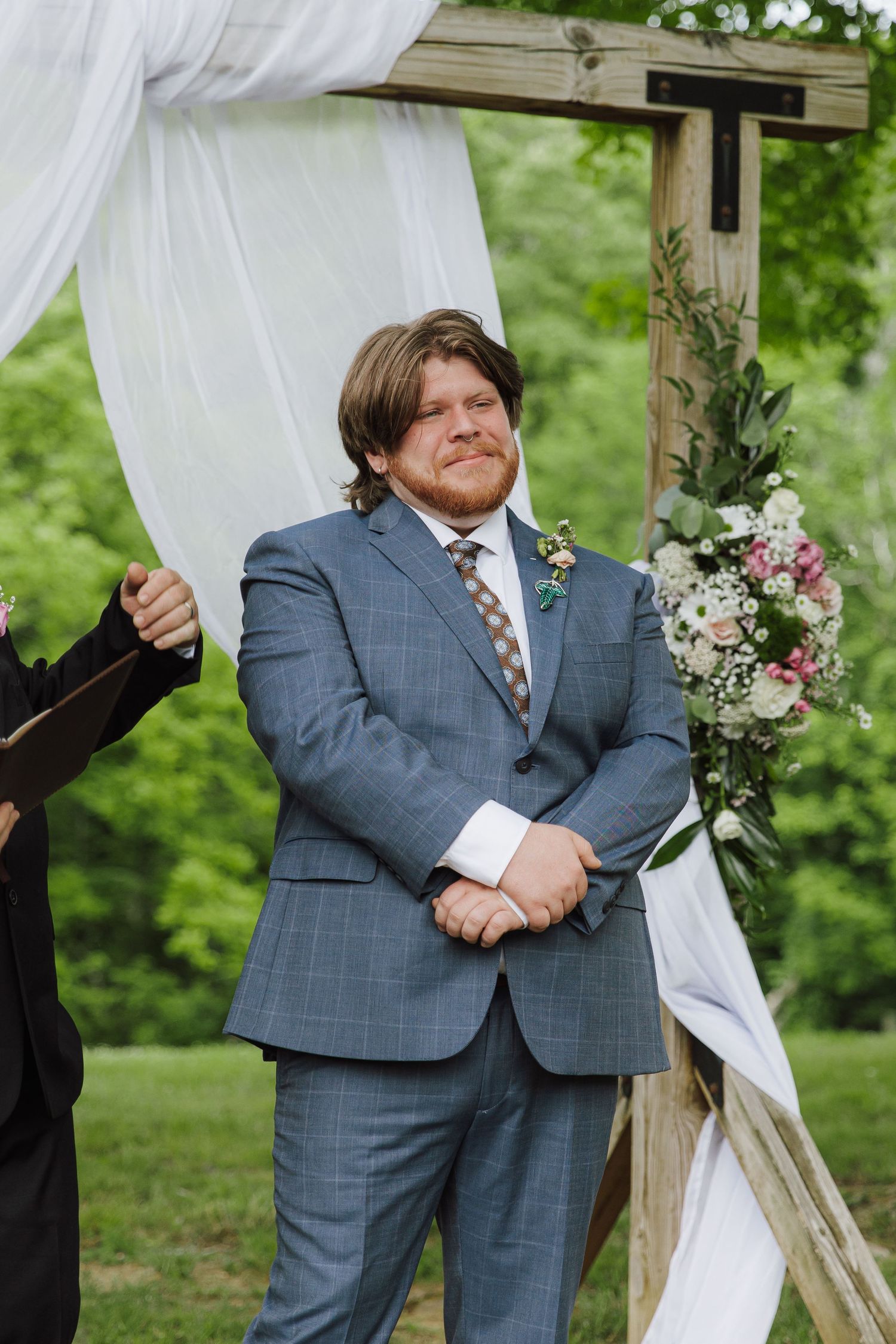 Emotional groom in blue plaid suit stands at floral wedding arch awaiting bride during outdoor ceremony.
