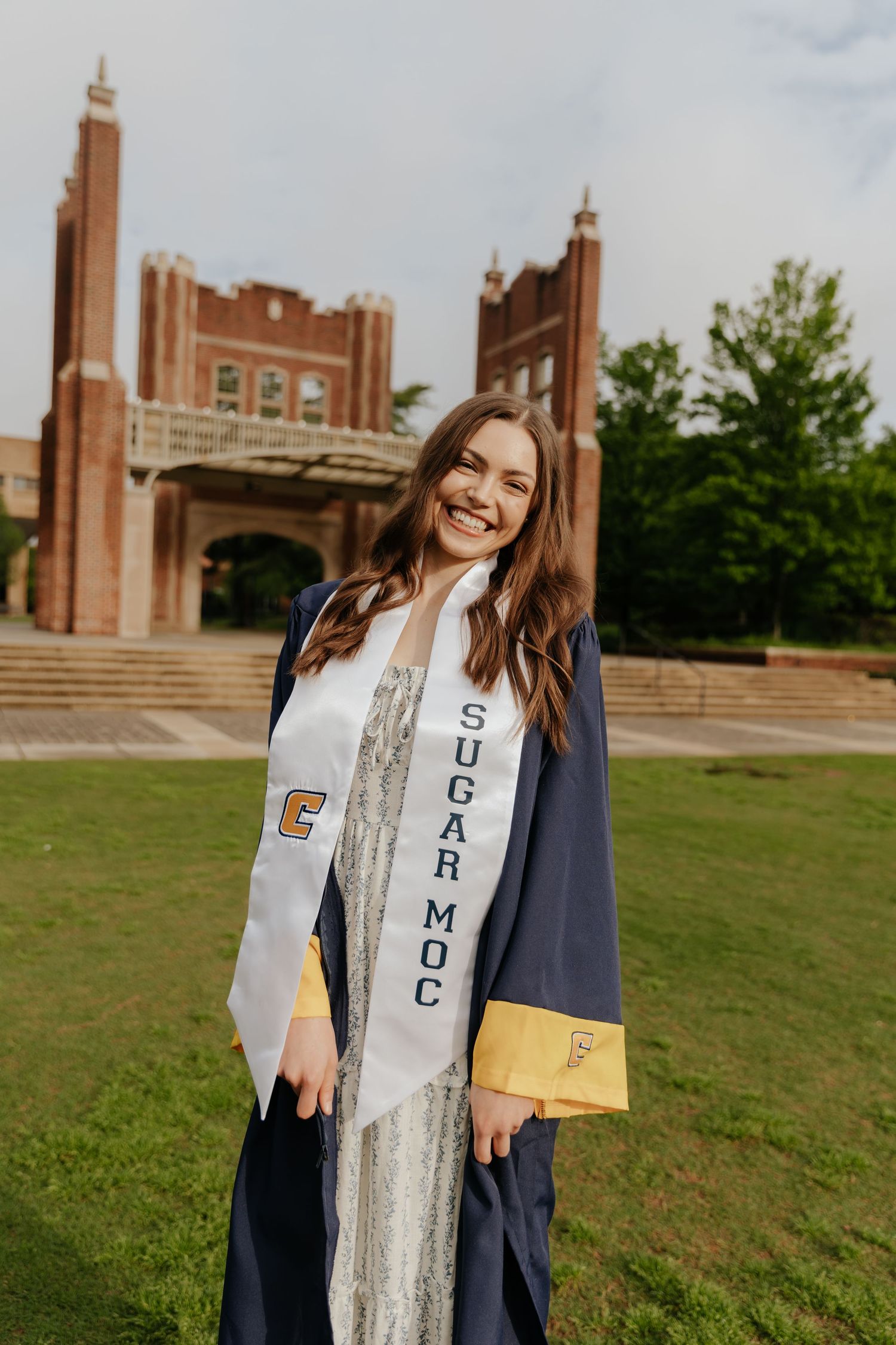 Graduate in blue and gold academic regalia stands smiling on campus lawn with brick university building in background.