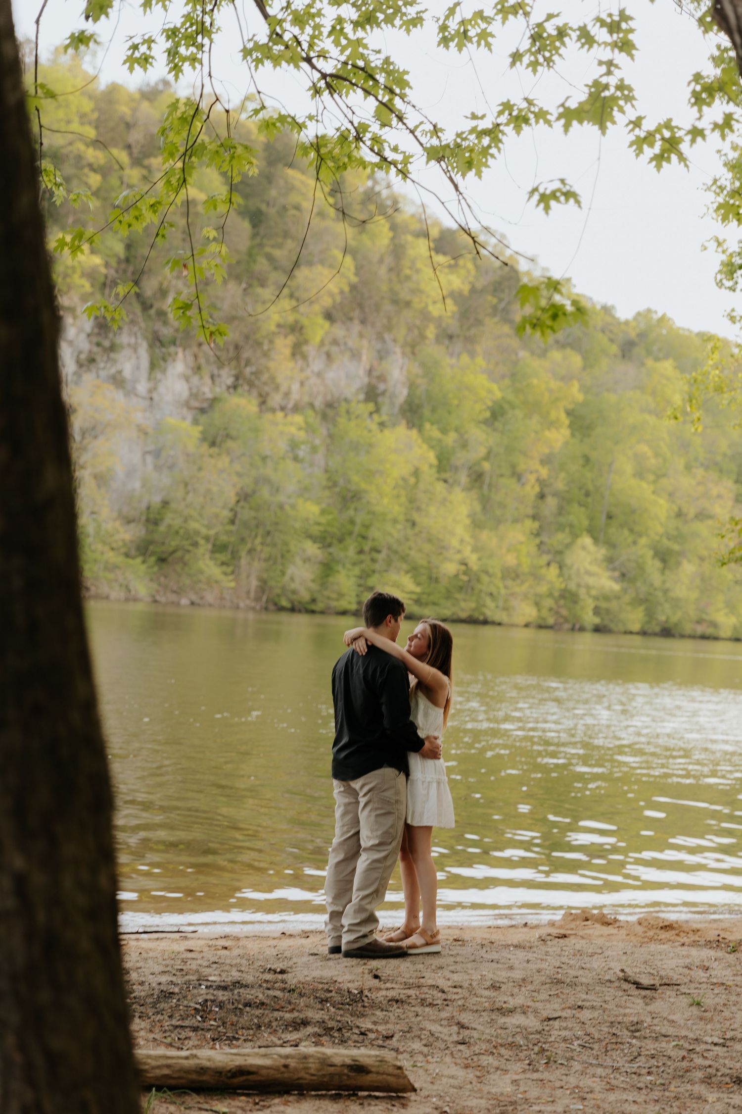 A romantic moment between two people standing by a serene lake surrounded by lush hillsides.