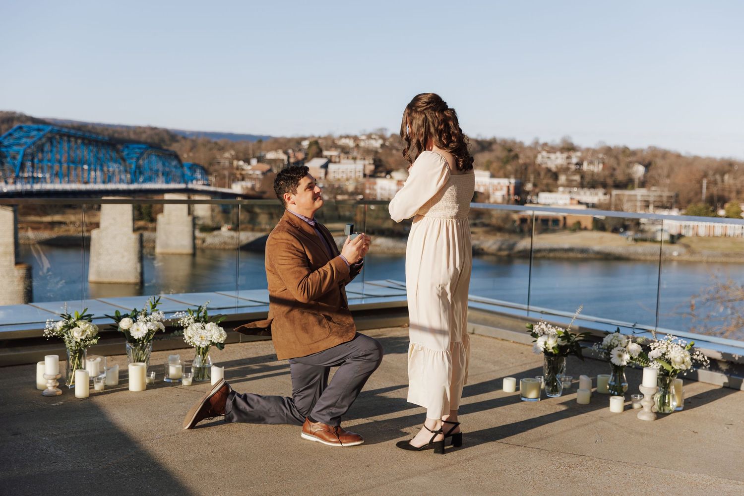 A romantic proposal scene with candles on a rooftop overlooking a river and bridge.