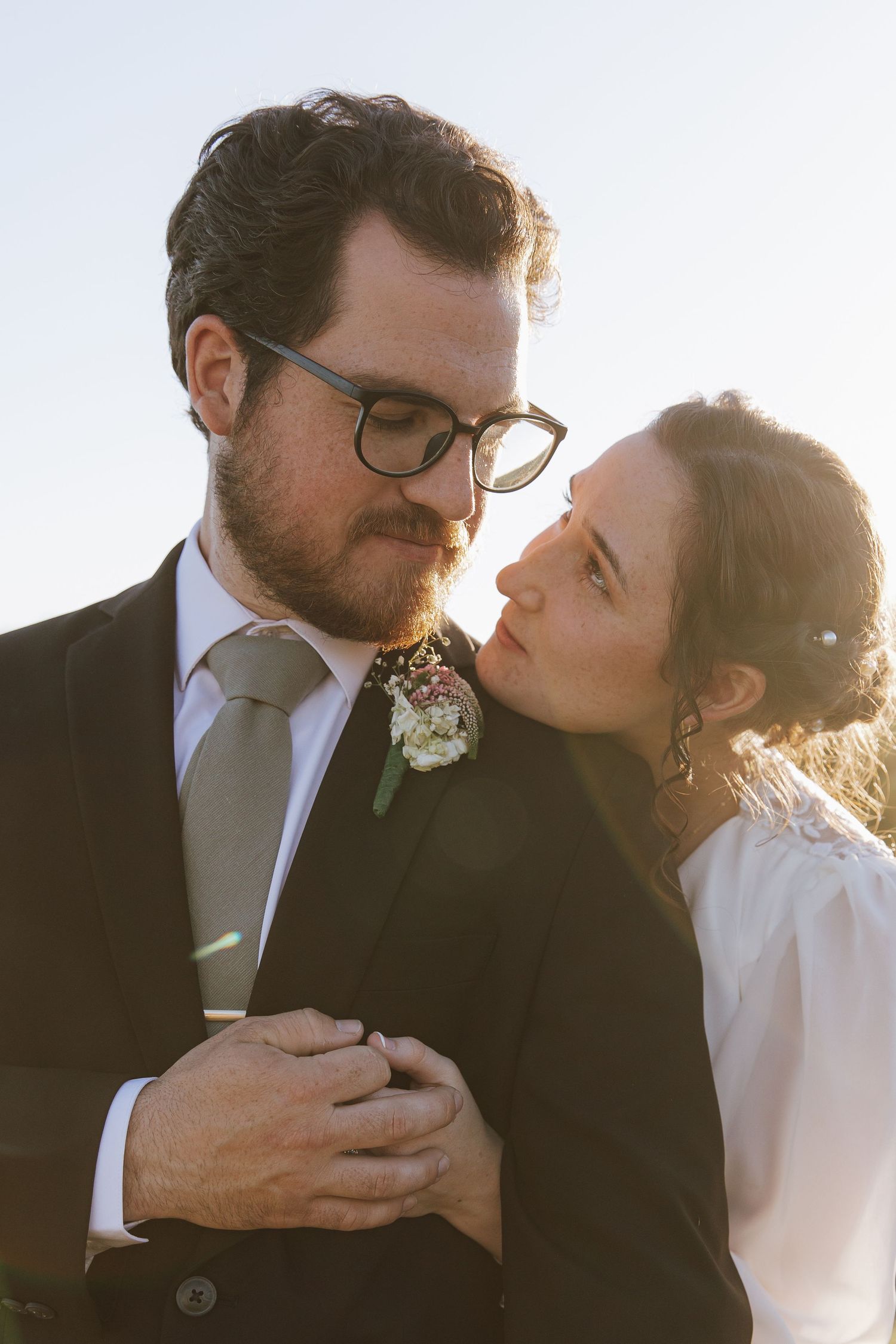 Close-up of a bearded groom in glasses and suit with boutonniere as bride embraces him at sunset.