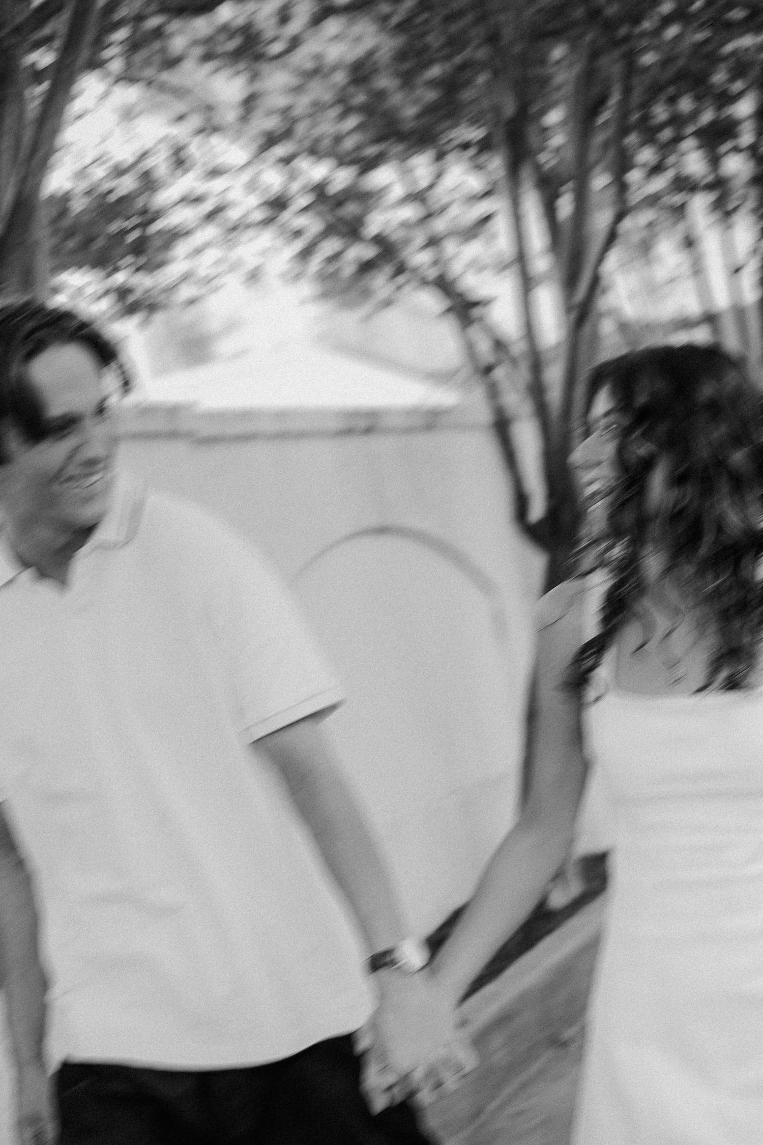 Black and white photo of a couple holding hands and smiling at each other outdoors near trees.