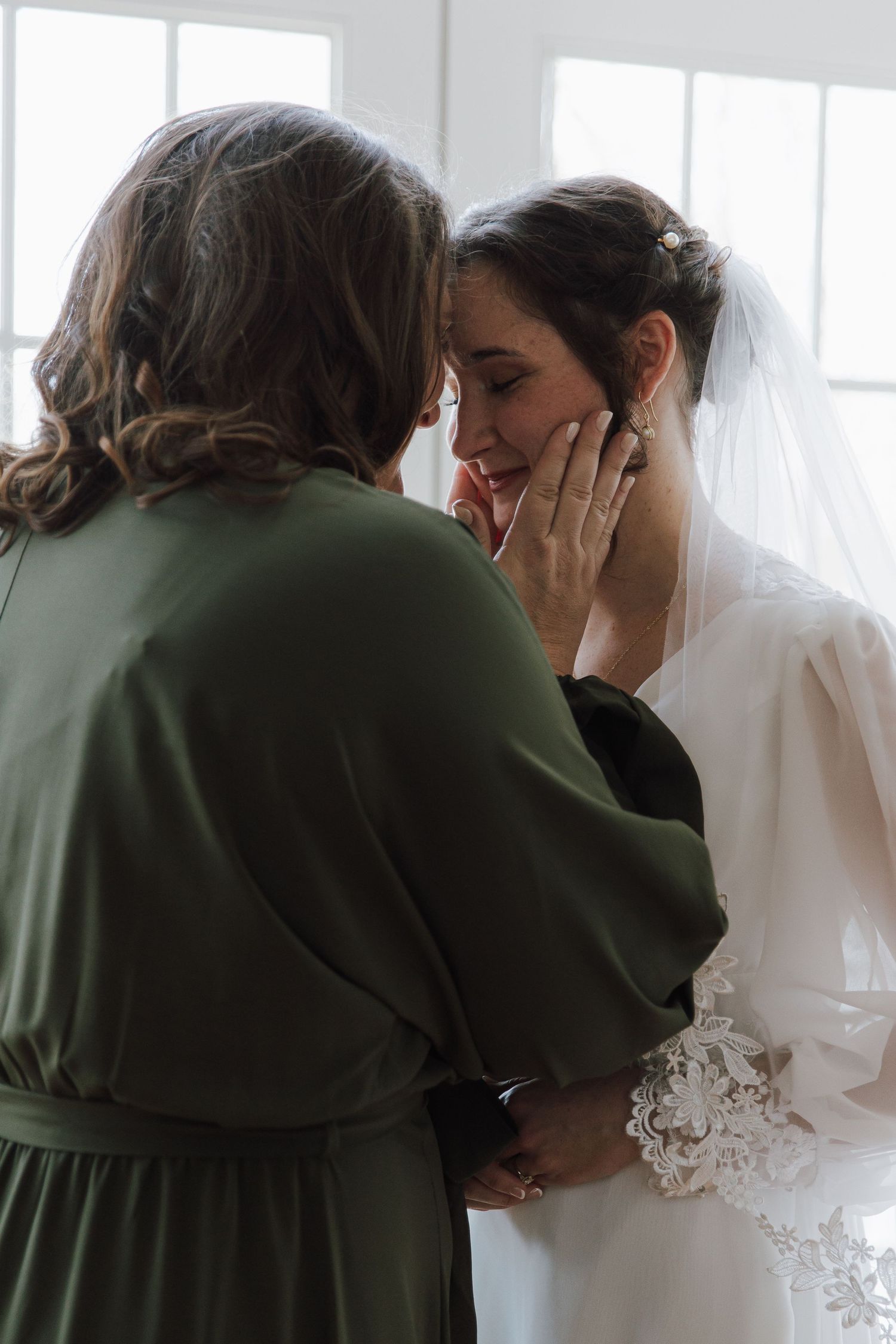 Tearful bride in lace veil being comforted by woman in olive green dress indoors.
