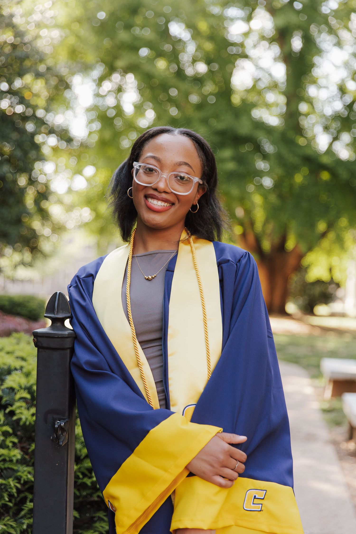 Smiling graduate in blue and gold cap and gown poses outdoors on campus.
