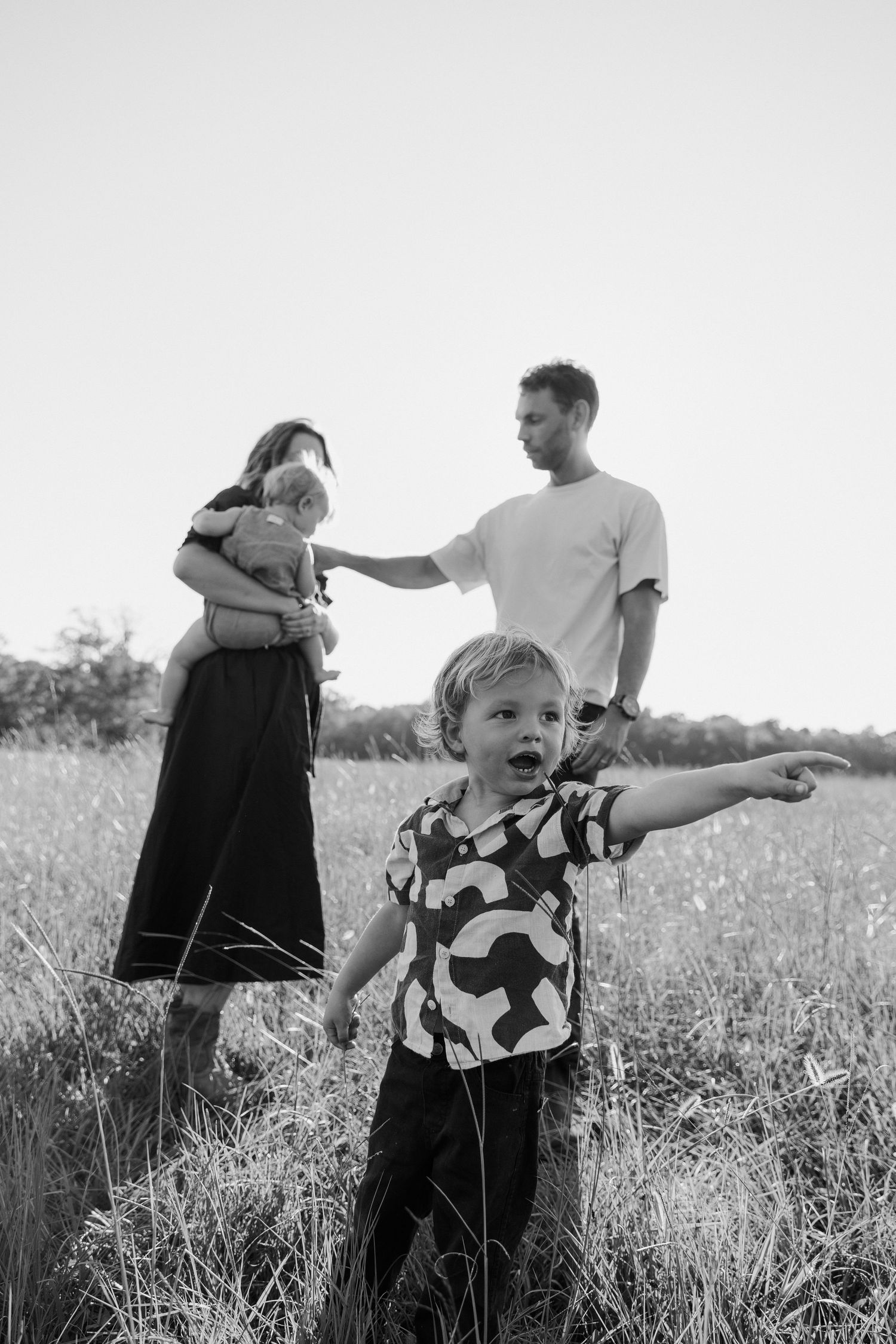 Black and white photo of a family outdoors in a grassy field with a child pointing excitedly in the foreground.