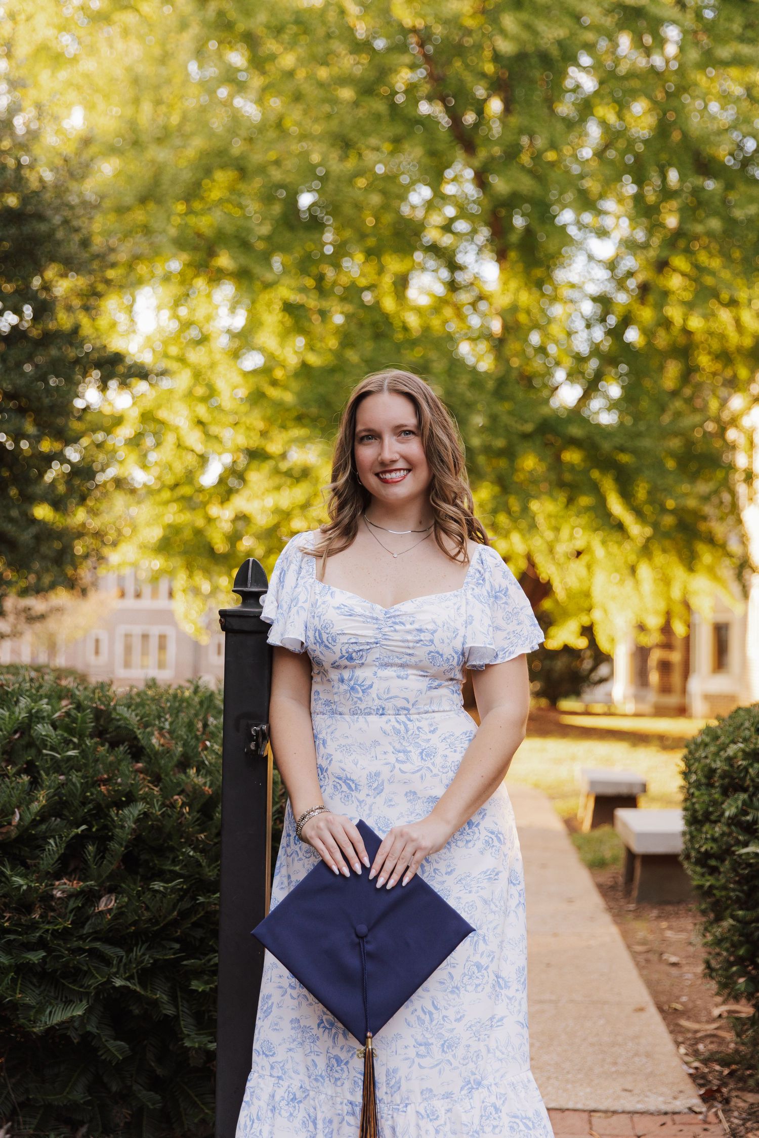 Graduate in a white and blue floral dress poses with diploma on campus during golden hour.