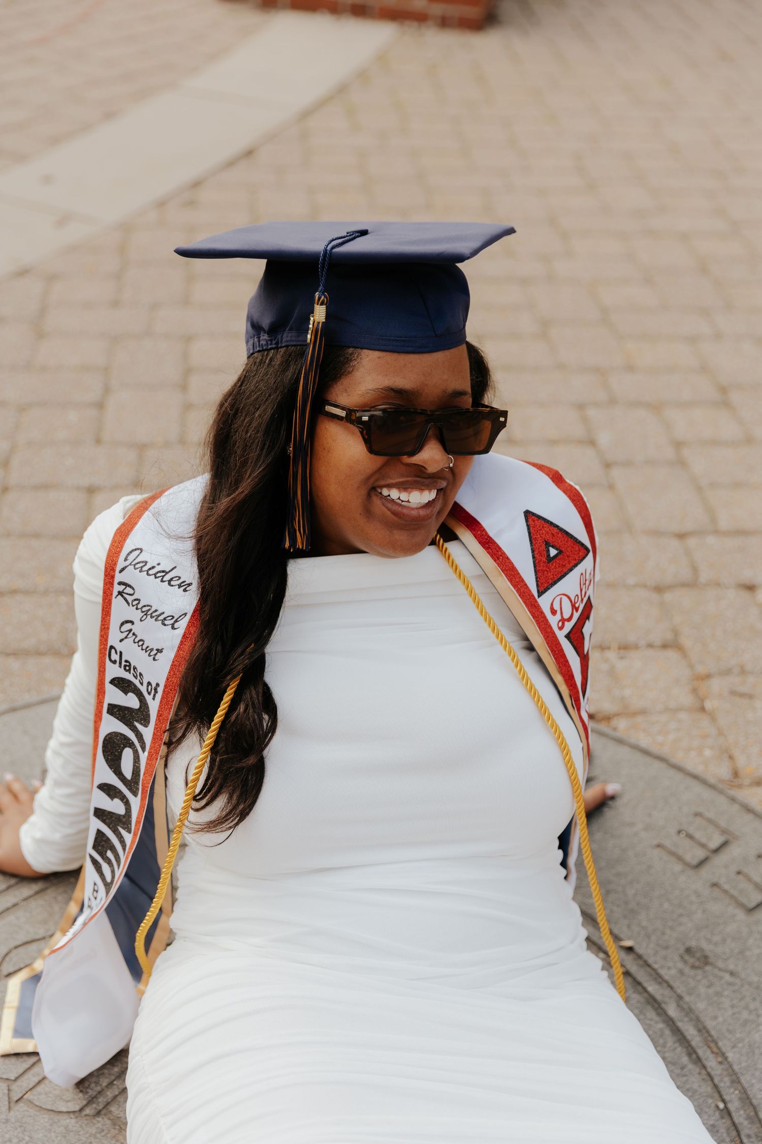 A graduate in sunglasses and white dress wears academic regalia and sorority stoles.