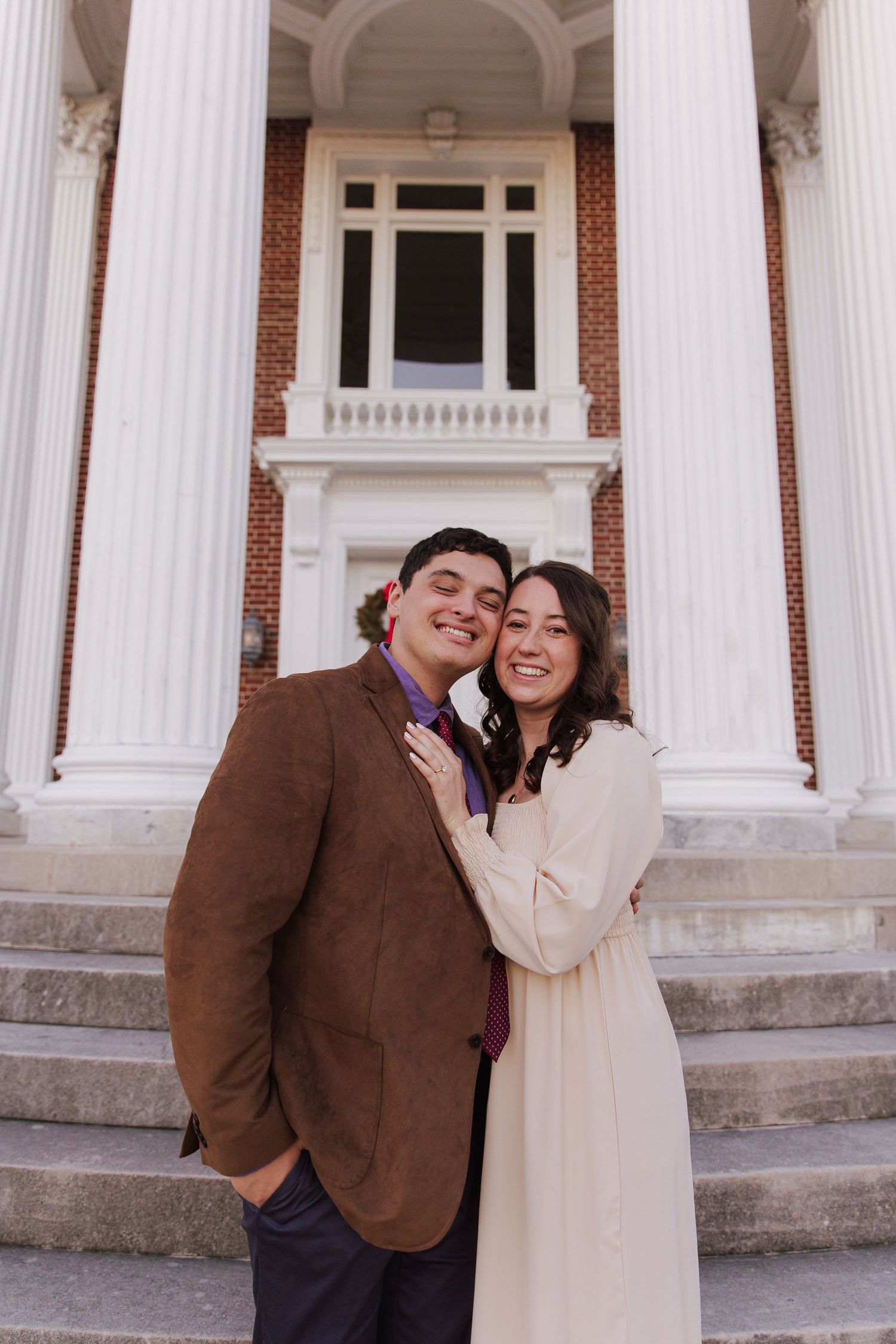 A couple laughs and embraces in front of classic white columns on a brick building.