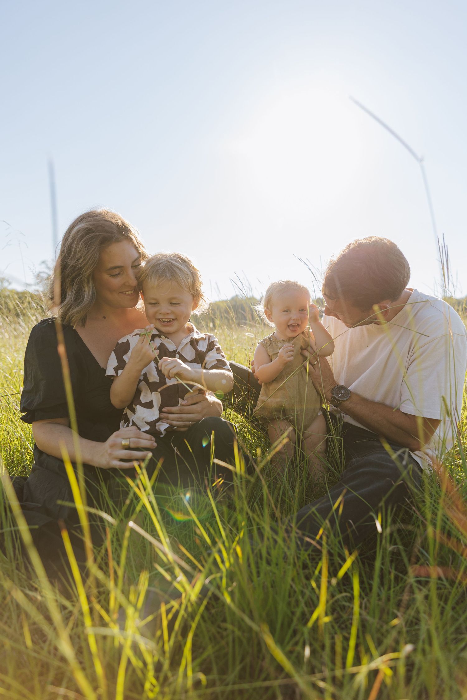 A family lies together in tall grass during golden hour, sharing tender moments in a sun-drenched meadow.