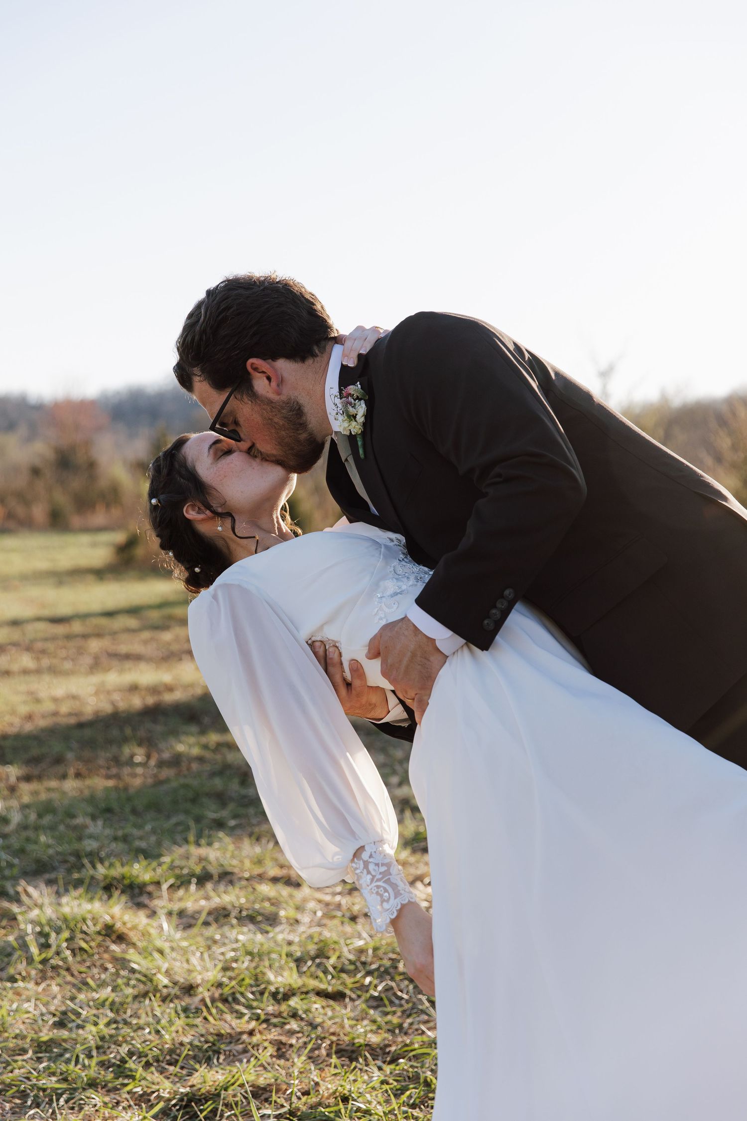 Groom dips bride for romantic kiss outdoors on sunny wedding day.