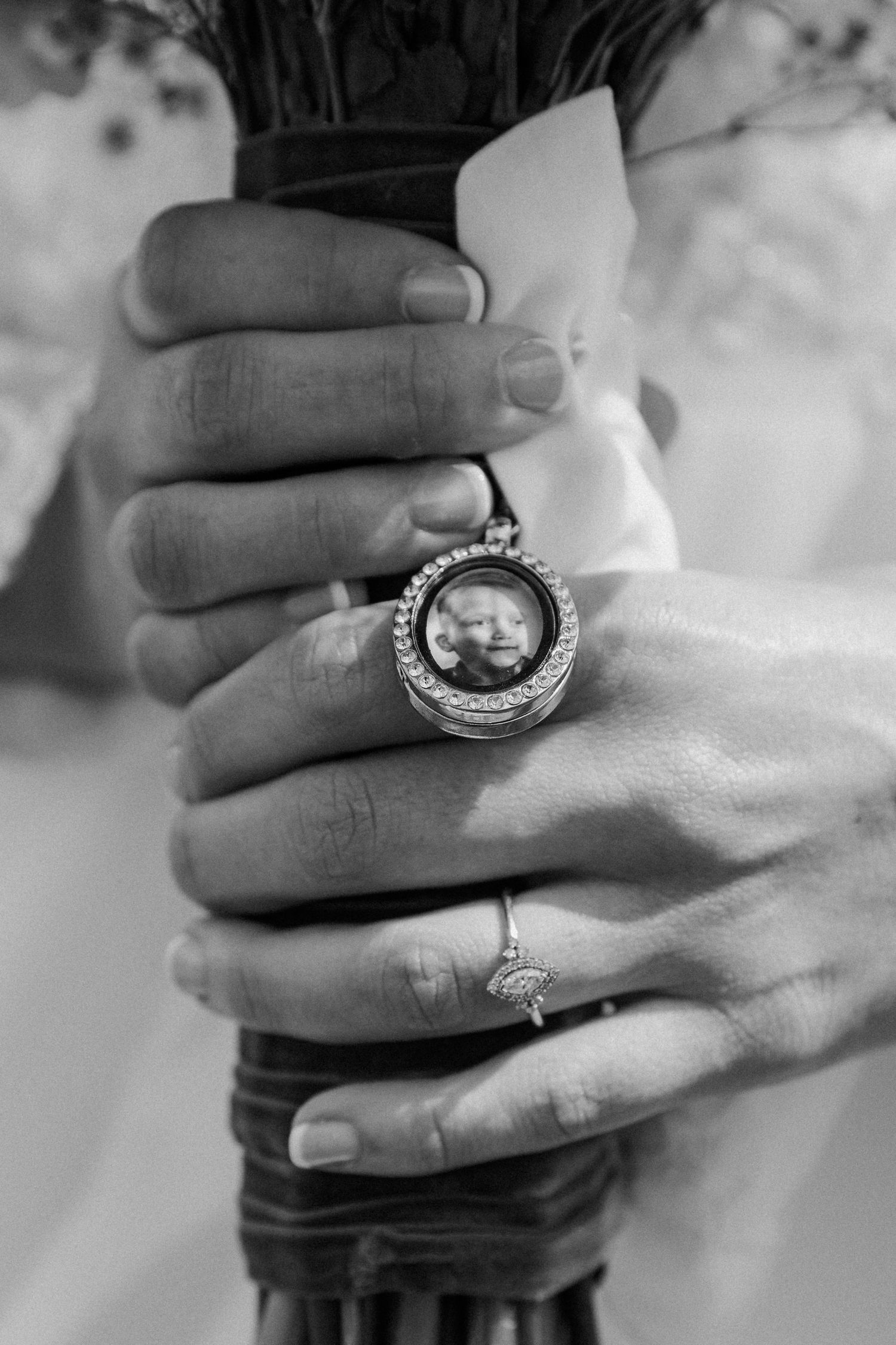 Close-up black and white photo of bride and groom hands with rings and locket bouquet charm.