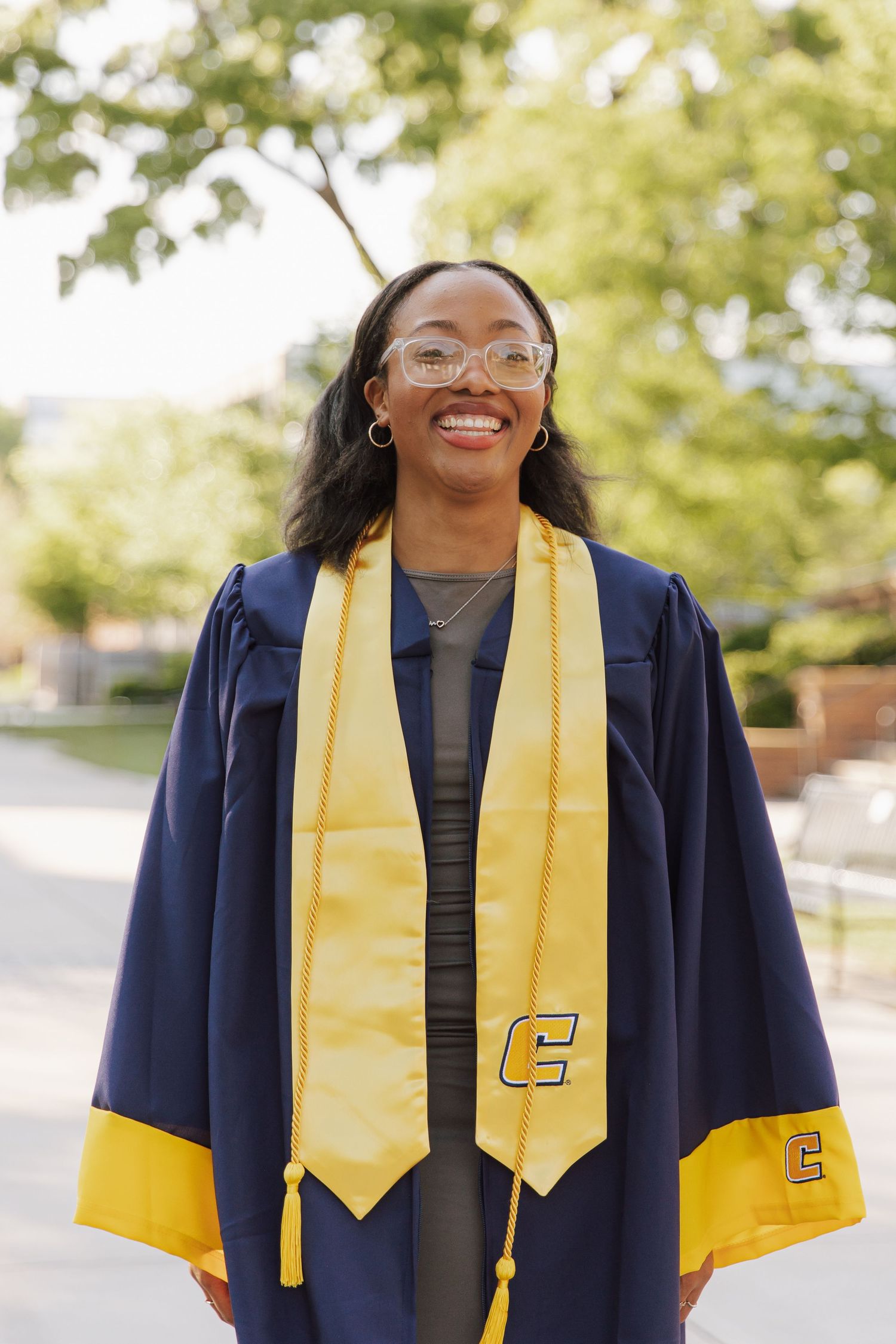 Smiling graduate in navy blue cap and gown with gold stole featuring letter C, wearing glasses outdoors.