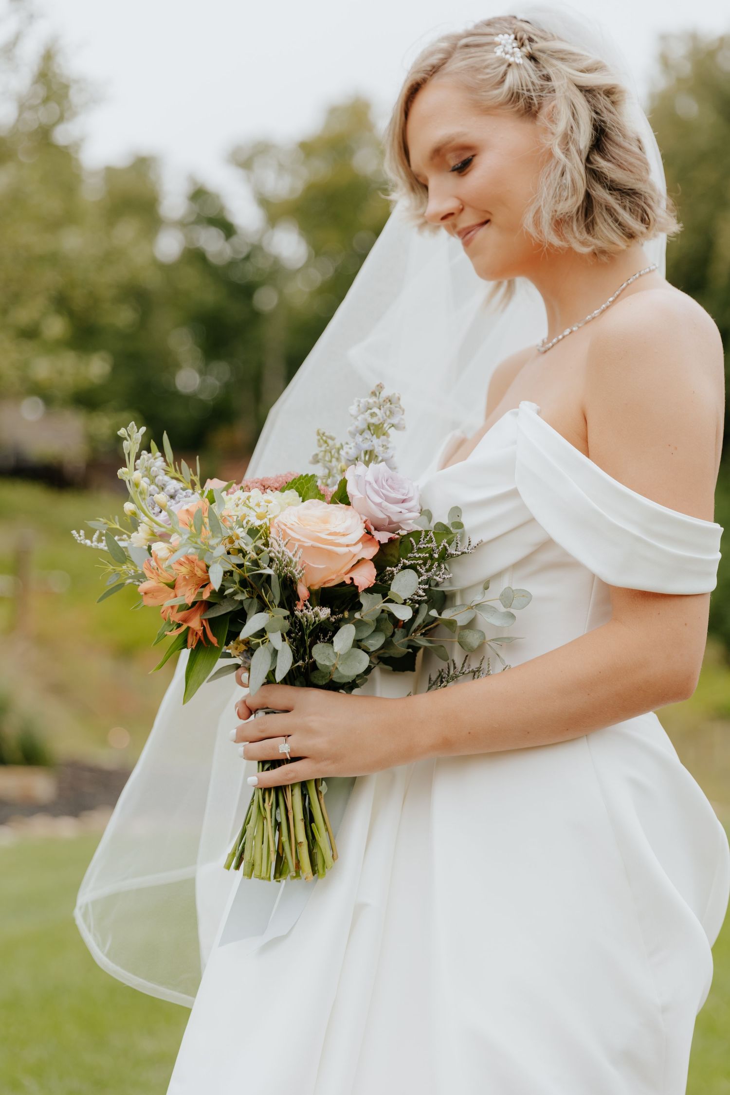 Bride in off-shoulder wedding dress holding colorful bouquet outdoors.