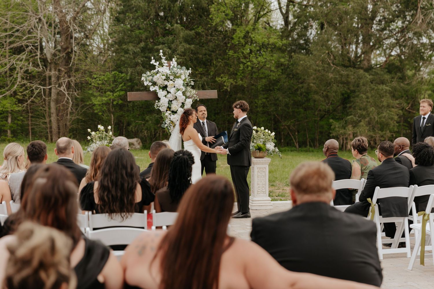 Wedding ceremony setup outdoors with white floral arch and chairs arranged on grass under trees.