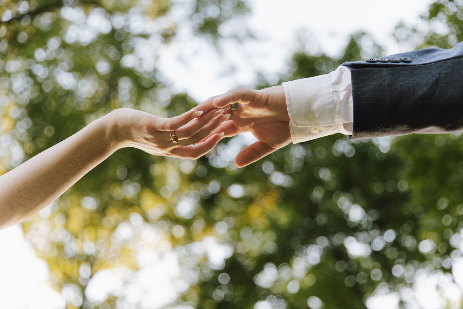 Two hands reaching toward each other tenderly against a soft green bokeh background, symbolizing a romantic wedding moment.