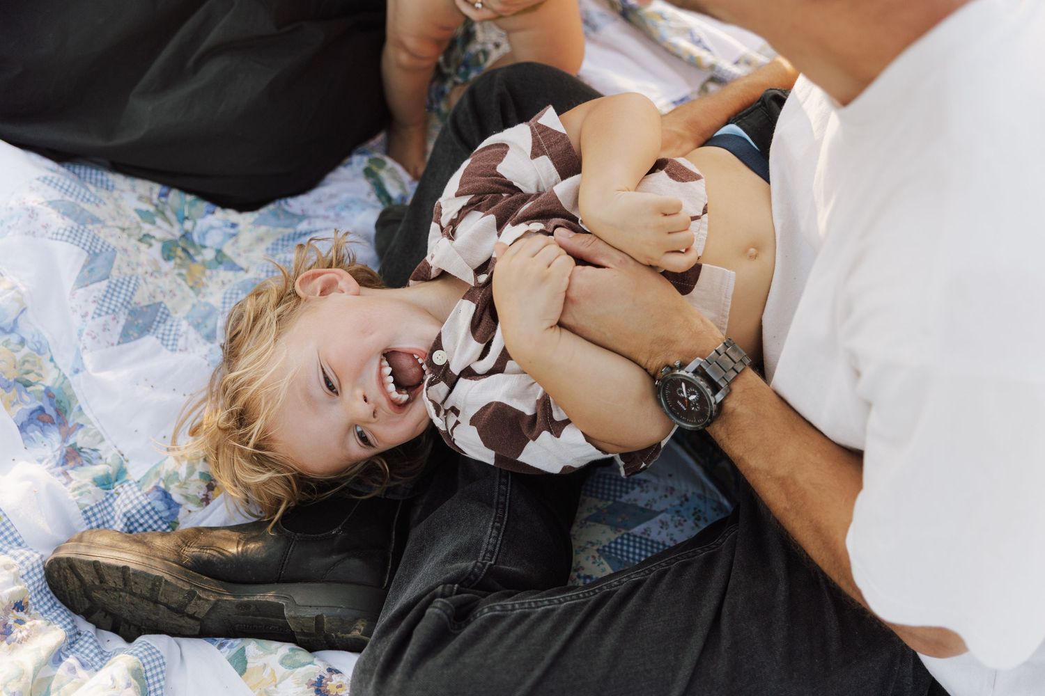 A young child laughs while being tickled on a blanket outdoors.