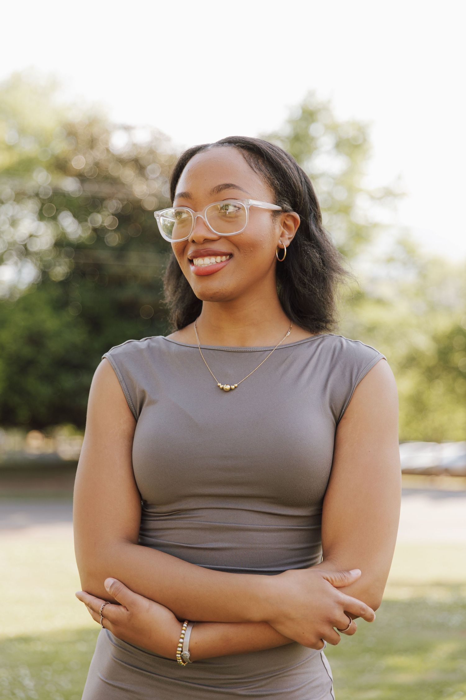Smiling young Black woman with glasses and gray dress stands outdoors with arms crossed, wearing gold jewelry.