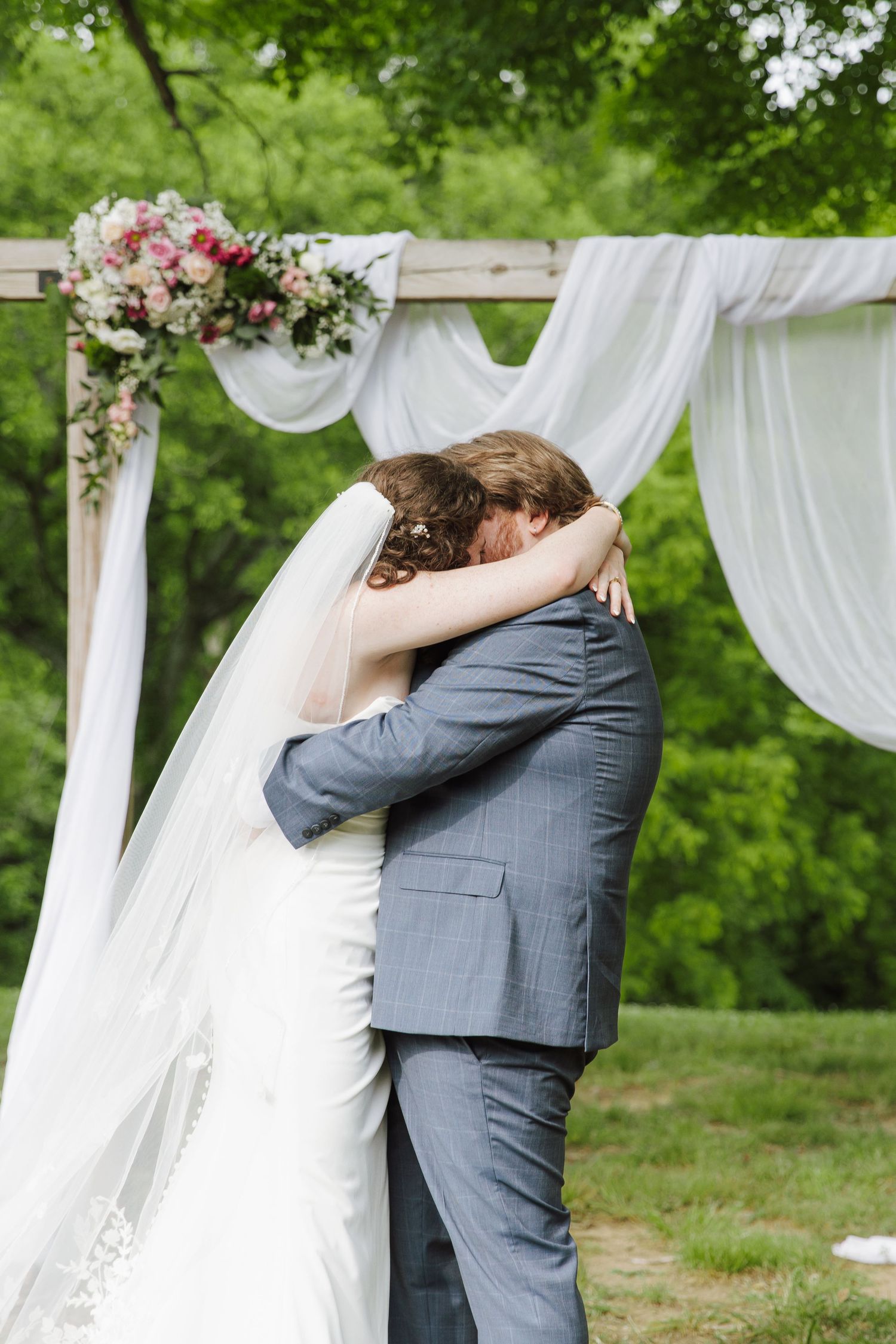 Bride and groom share first embrace under decorated wooden arch with white draping at outdoor wedding.