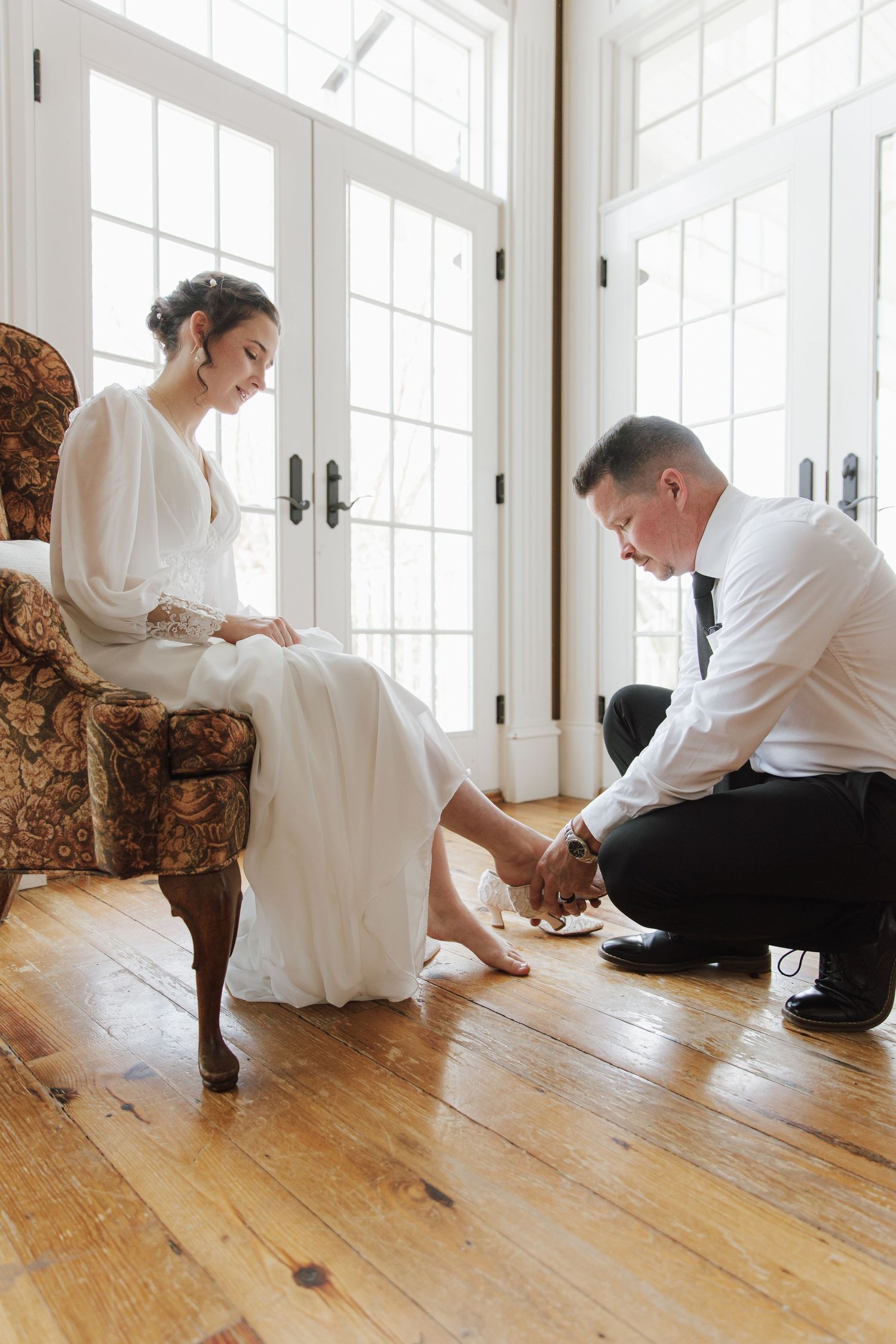 Groom kneeling to put shoes on bride sitting in ornate chair by French windows on wedding day.