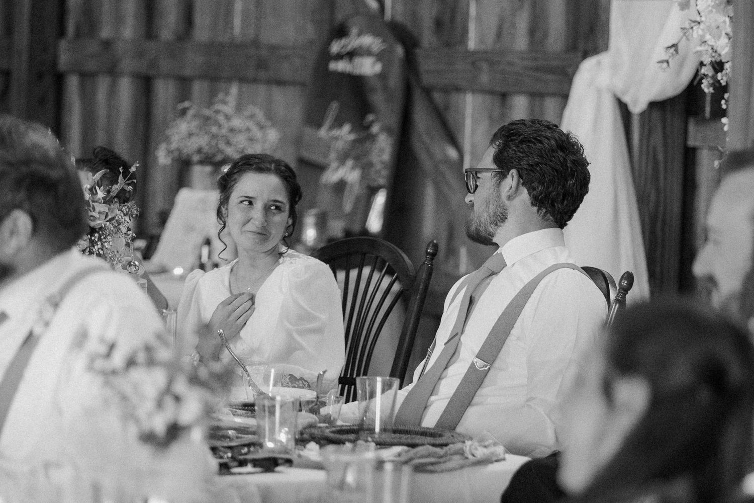 Black and white wedding reception photo of bride and groom seated at head table, sharing a joyful moment.