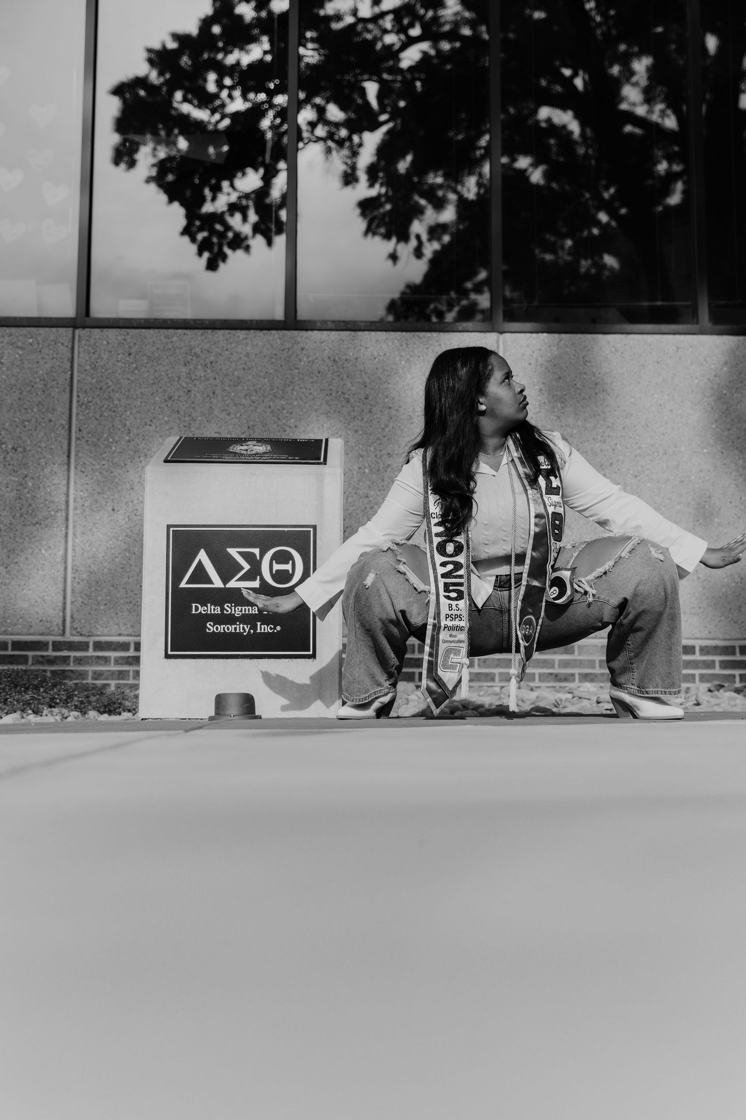 A graduate in casual attire poses near a Delta Sigma Theta sign in black and white photography.