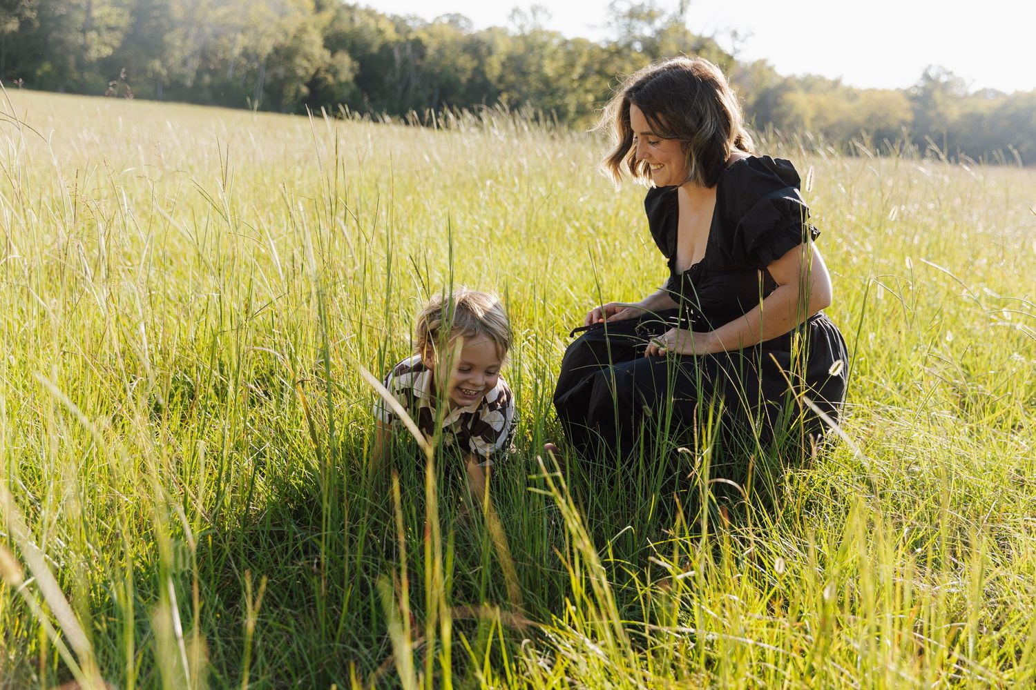 A mom and her son sit together in a sunlit meadow of tall grass during golden hour.