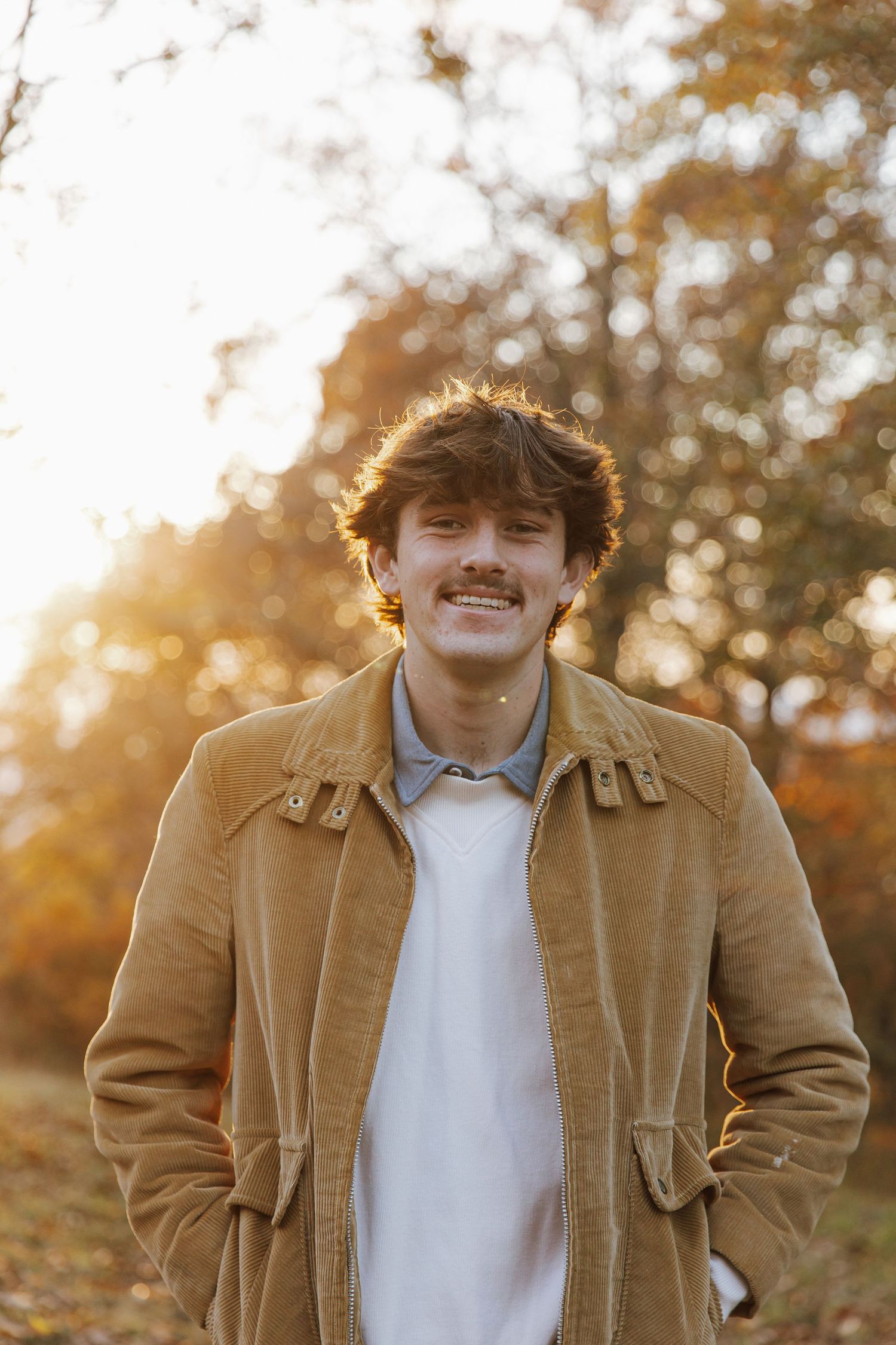 High school graduate in a brown corduroy jacket stands in autumn sunlight with golden trees in the background.
