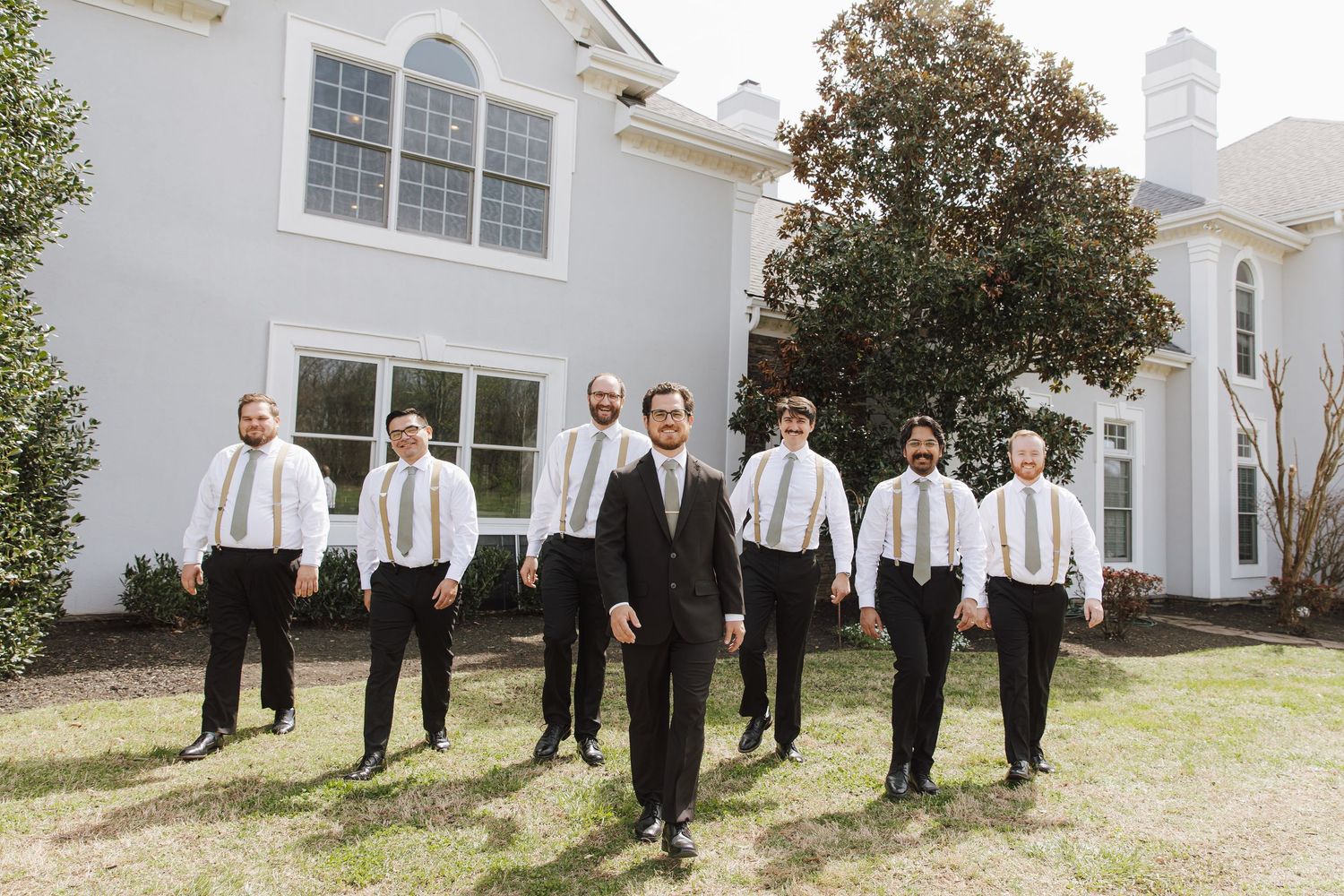Groom and groomsmen in black suits with gold ties walking outdoors in front of a white house.