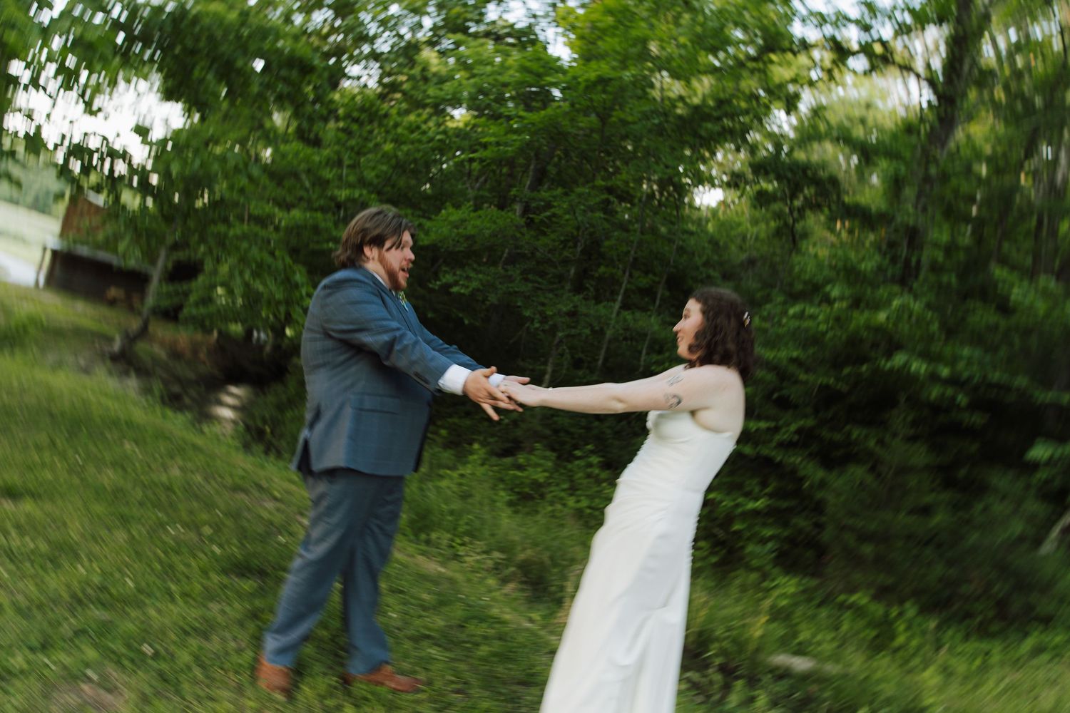 A bride and groom hold hands and dance together outdoors surrounded by lush green trees on their wedding day.