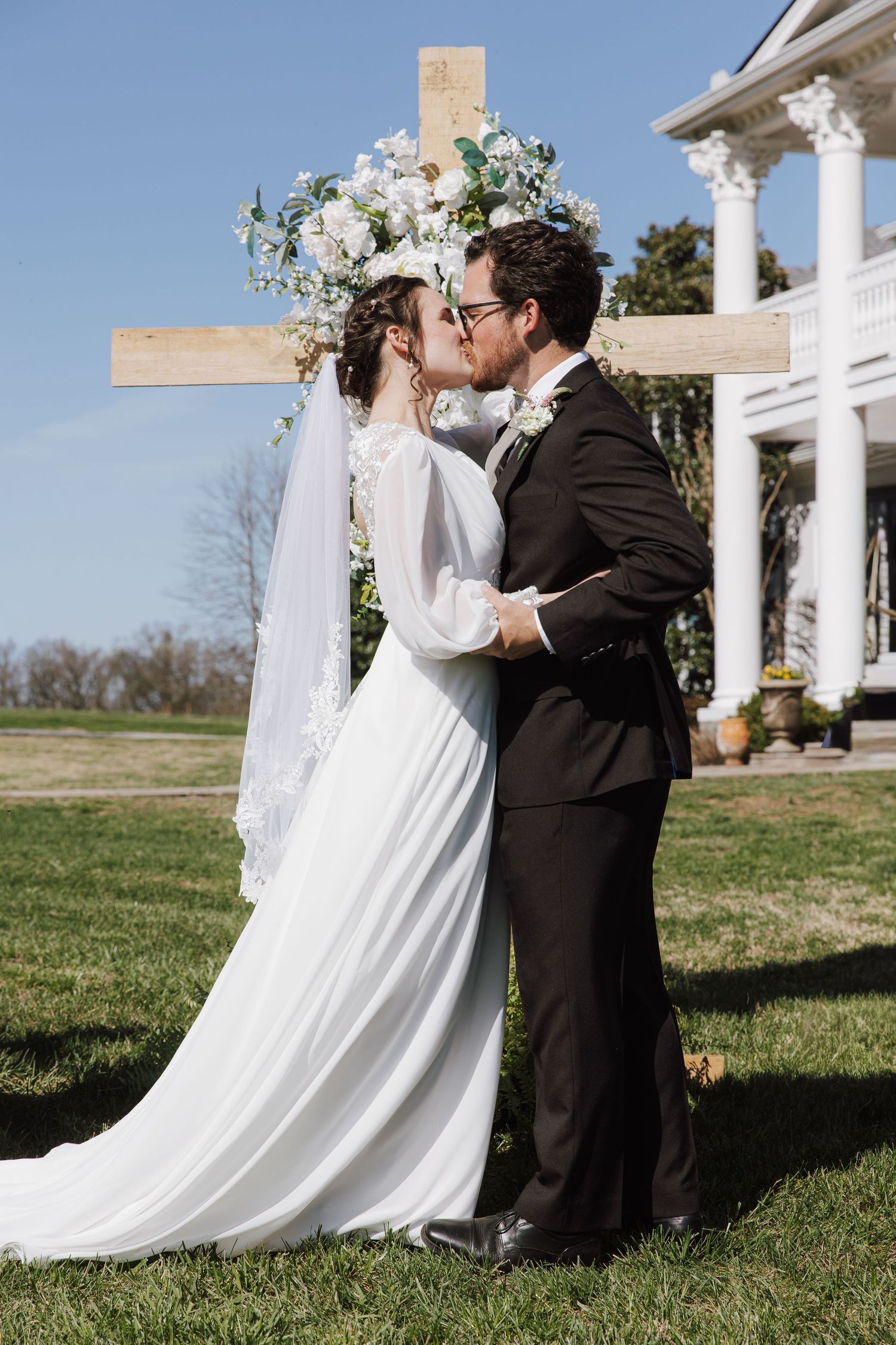 A romantic outdoor wedding ceremony scene with a white gown and black suit against a blue sky.