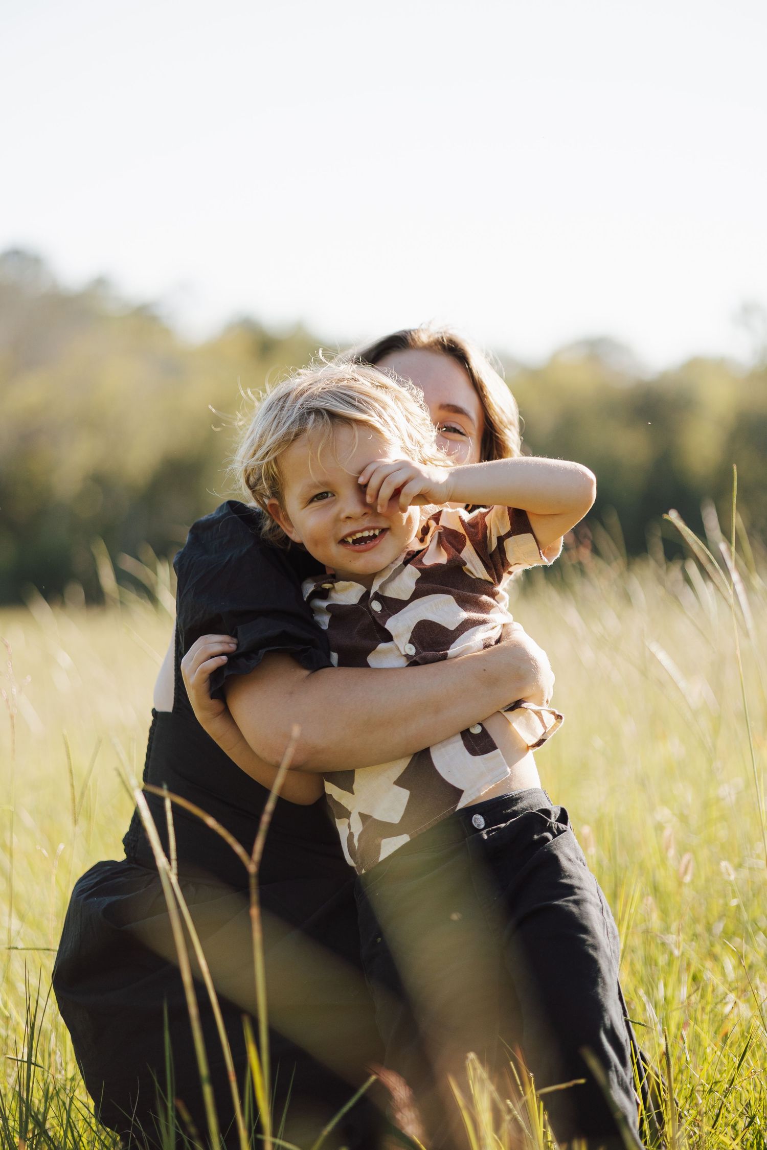 A mom hugging her young son from behind while he is looking at the camera.