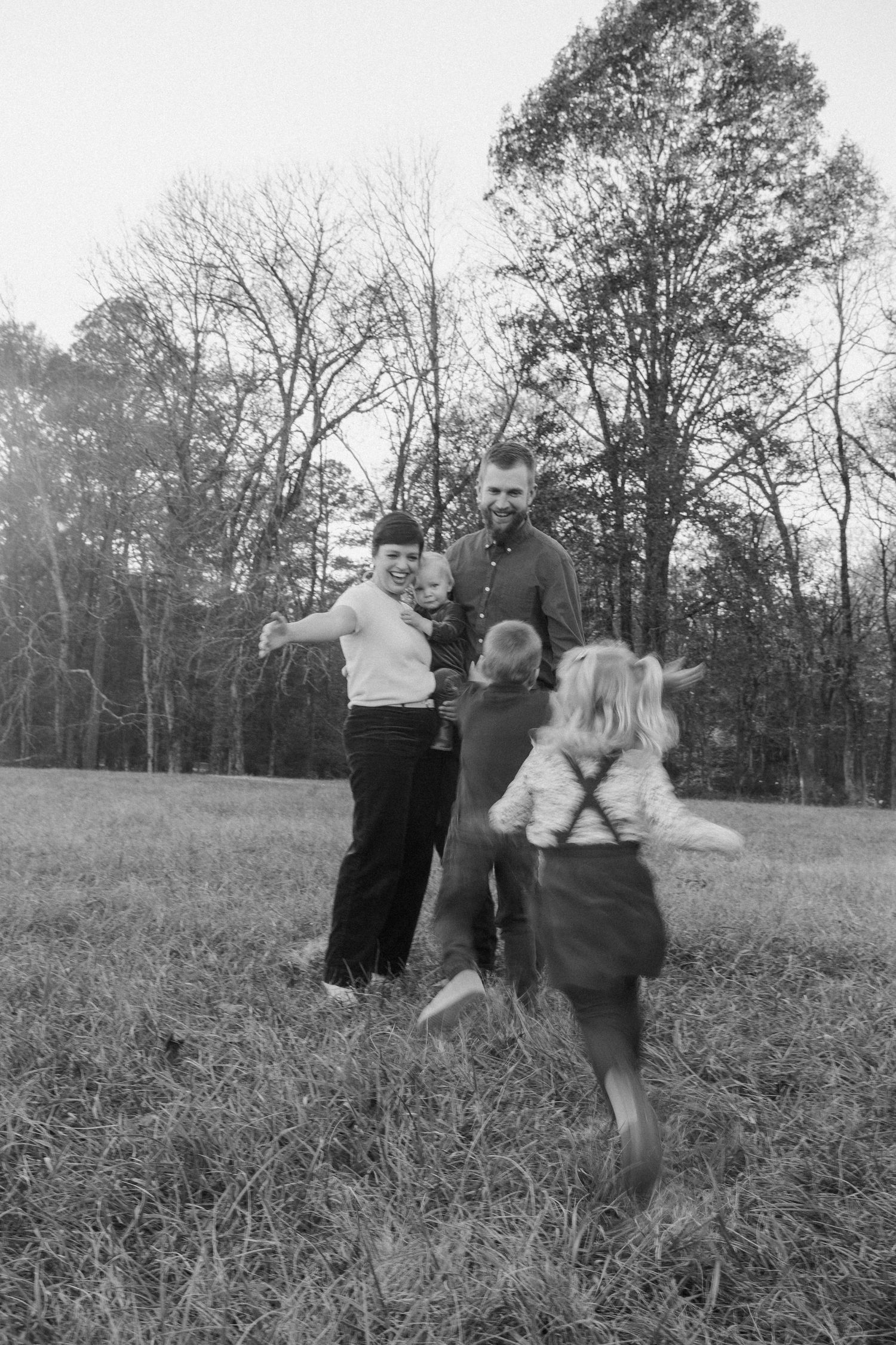 Black and white photo of a family playing together in an open field surrounded by bare trees.