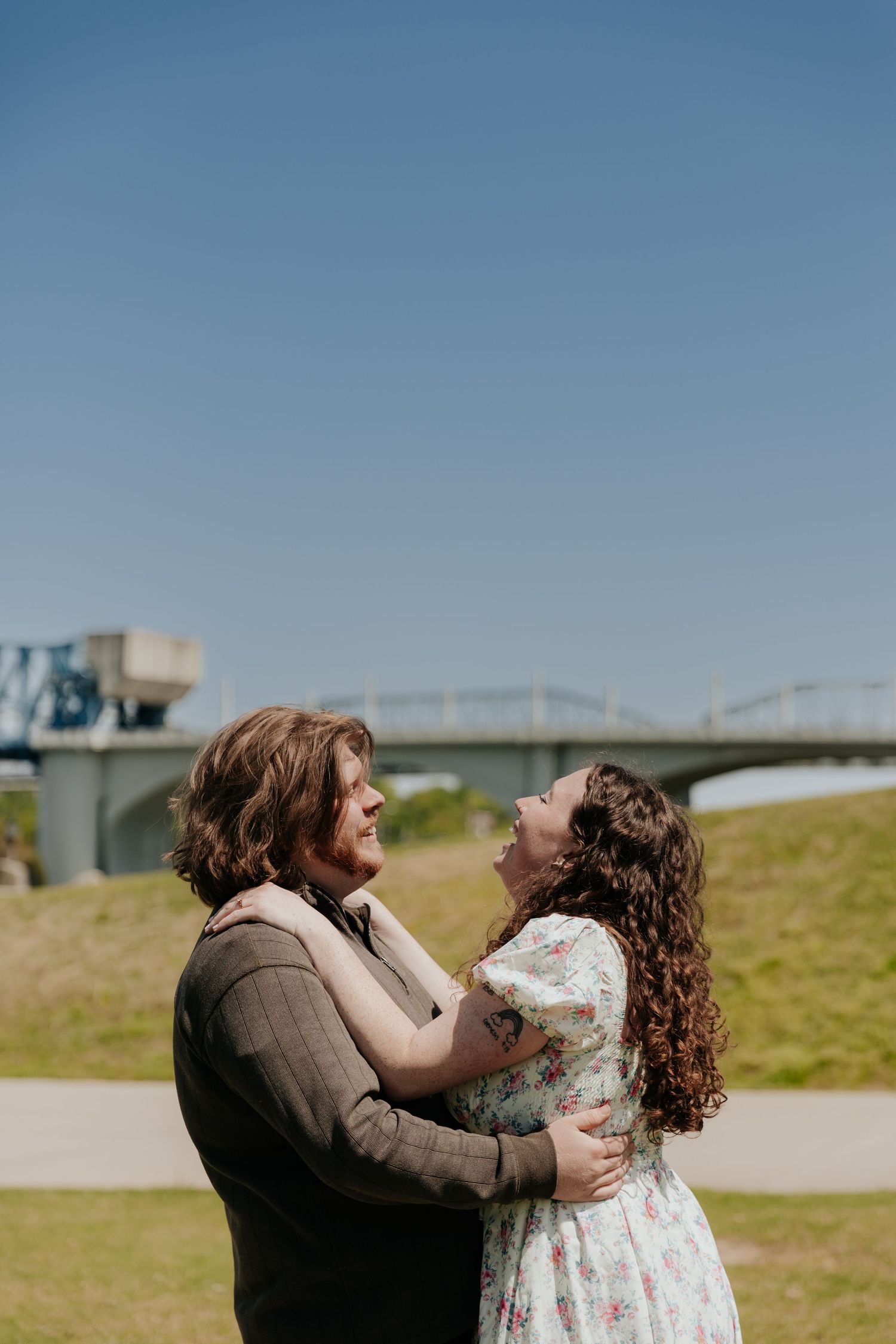 A joyful embrace captured on a sunny day with a bridge and green hillside in view.