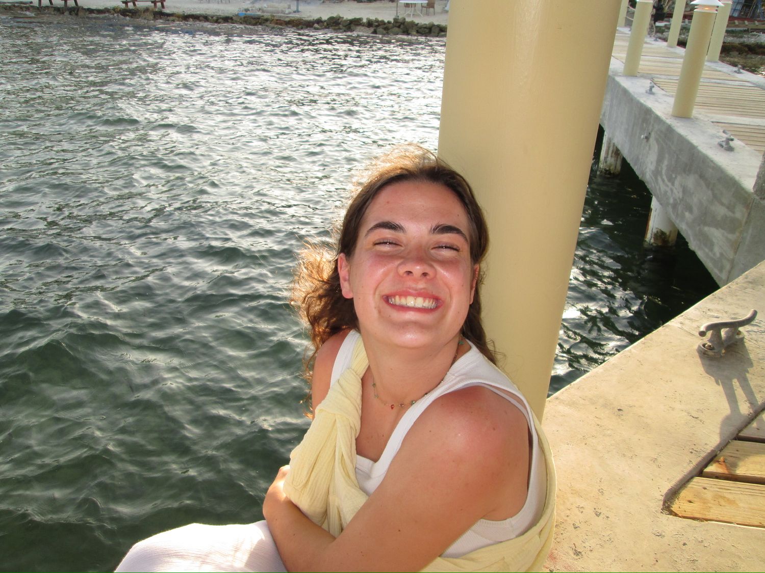 Someone smiling and laughing while sitting by a waterfront dock on a sunny day.