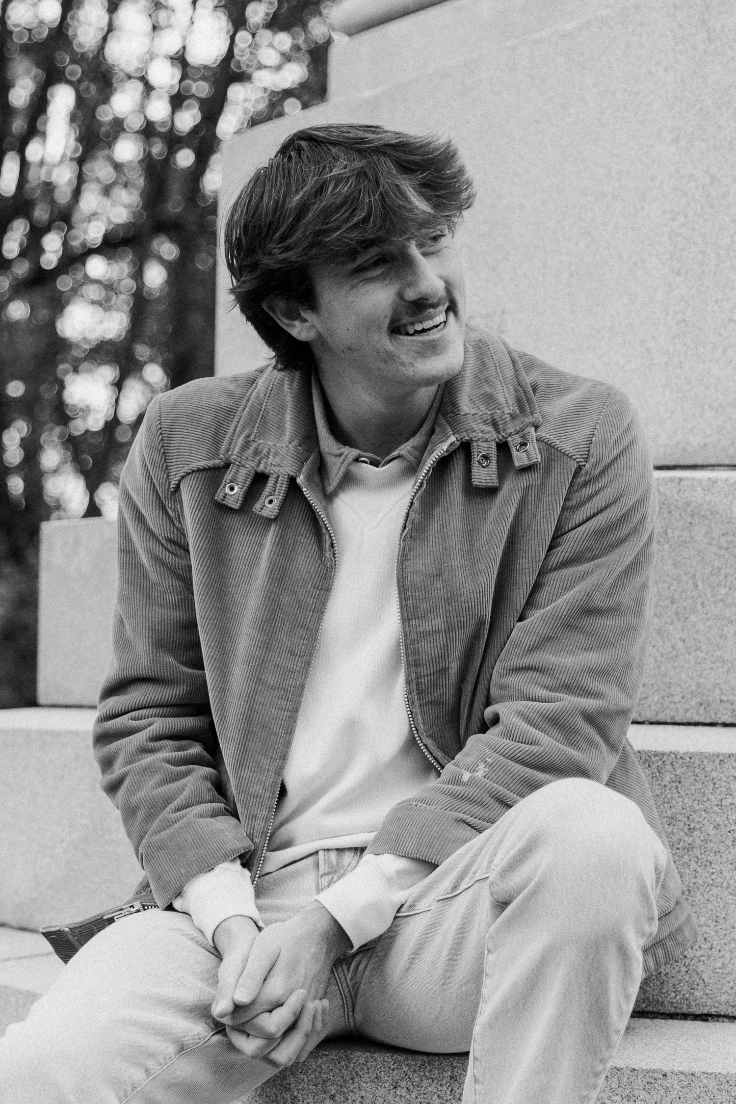 Black and white portrait of a smiling young person wearing a casual jacket while sitting on stone steps outdoors.