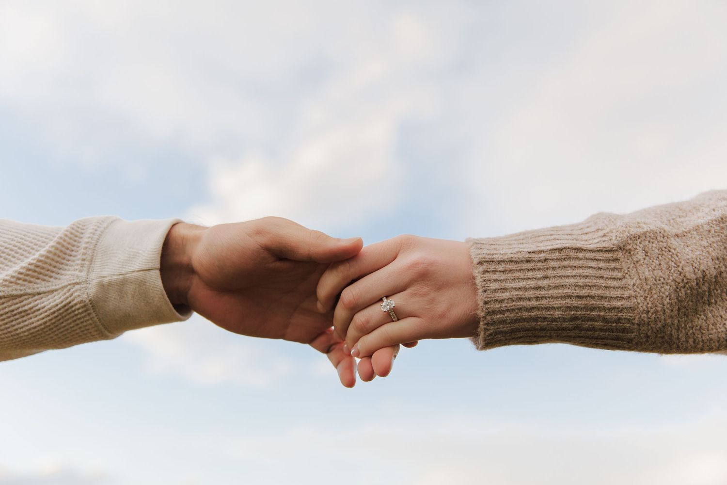 Two hands gently touching against a bright sky, showing an engagement ring.