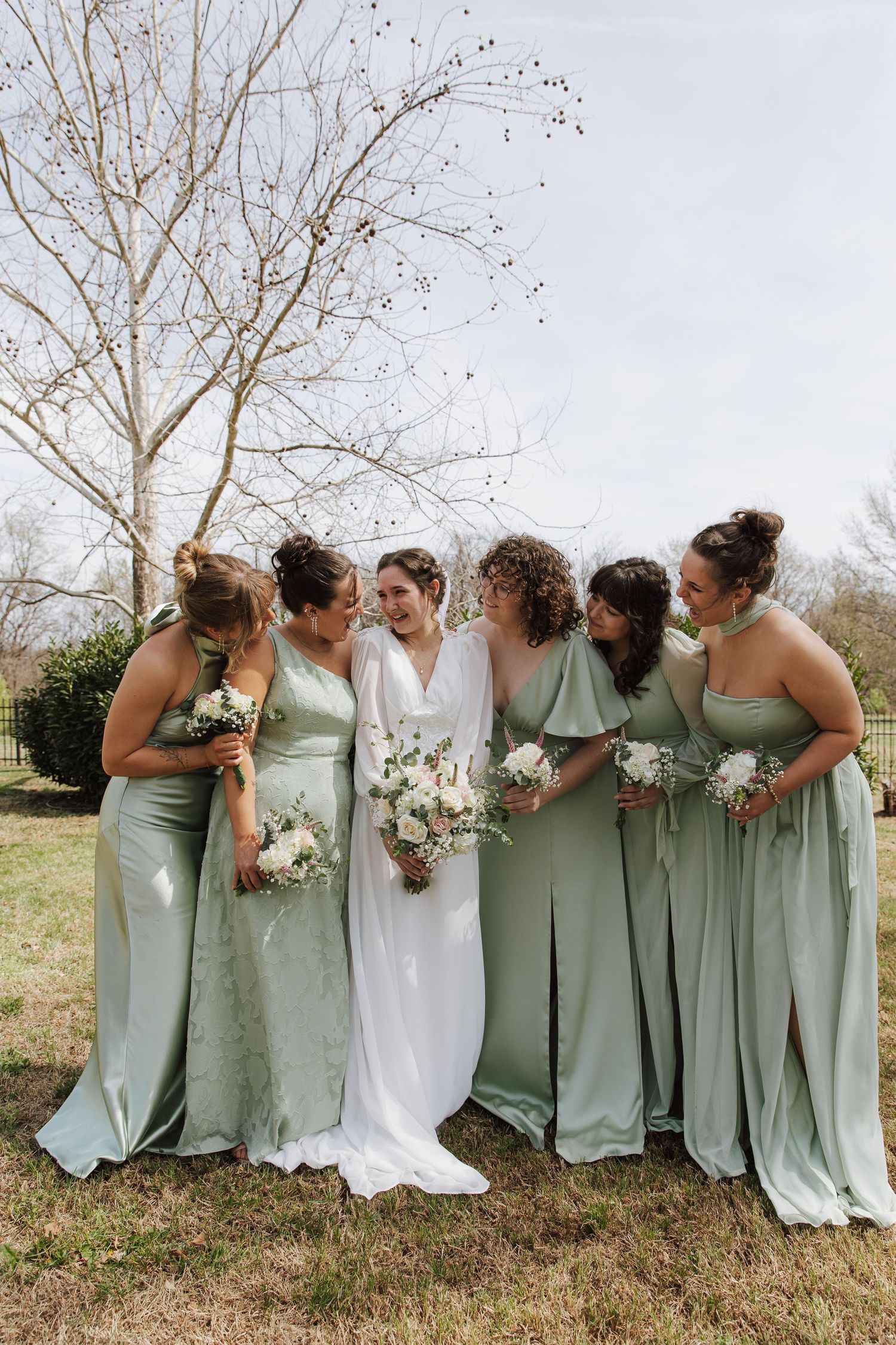 Bride in white gown with five bridesmaids in sage green dresses holding bouquets outdoors.