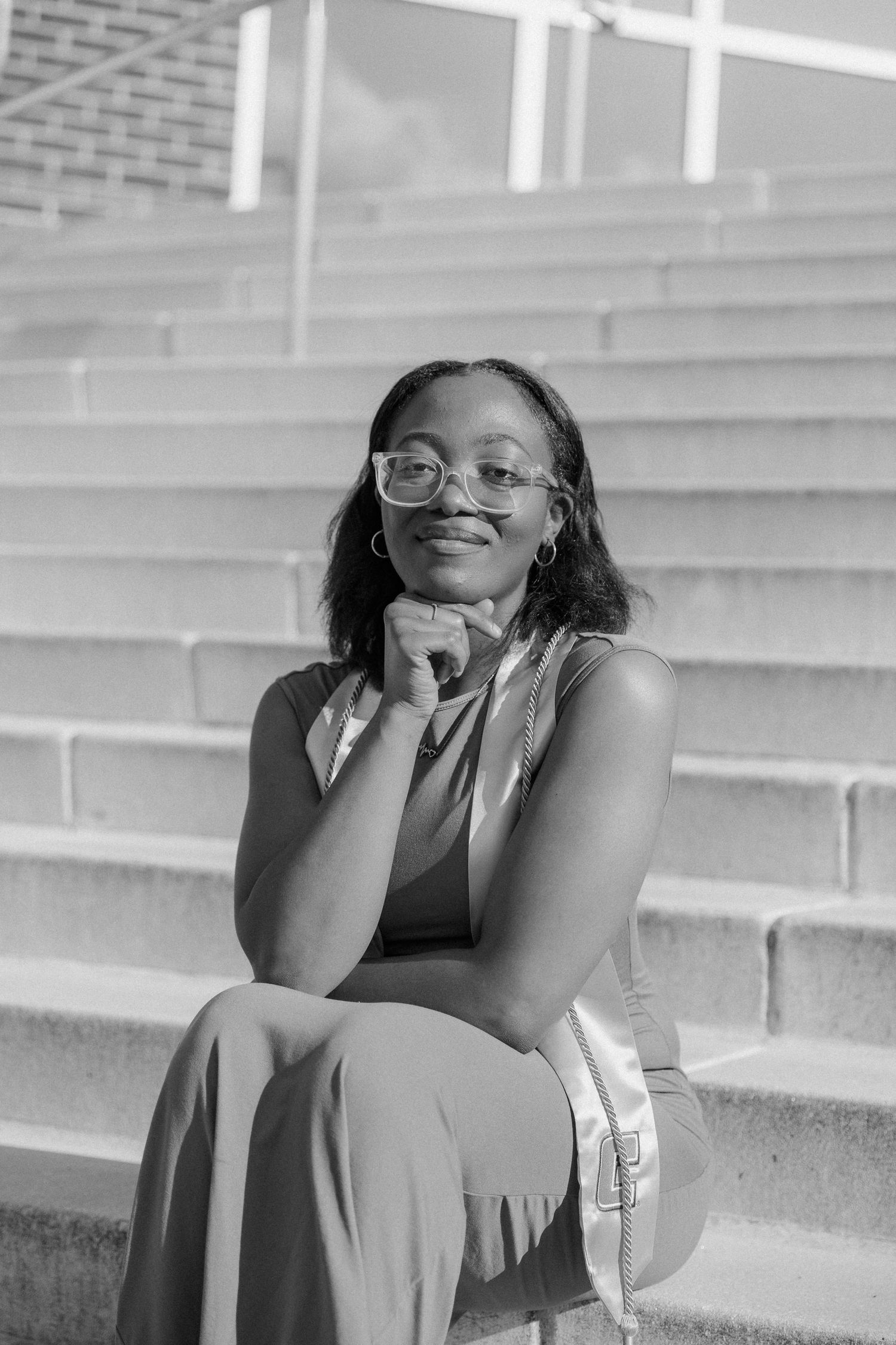 Black and white portrait of a young woman with glasses sitting on outdoor steps, smiling with hand on chin.