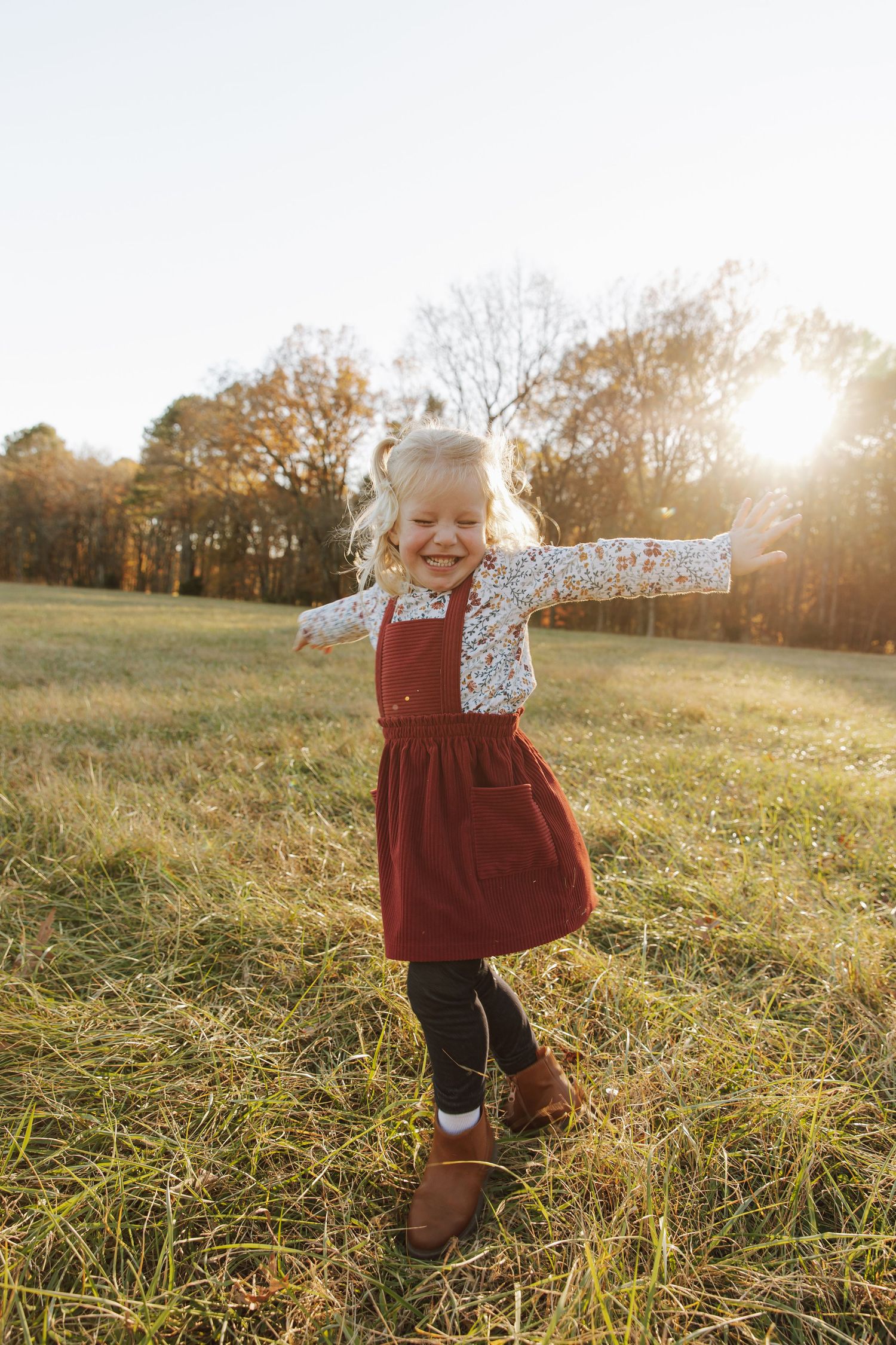 Young child in a burgundy dress twirling joyfully in a sunlit autumn field.