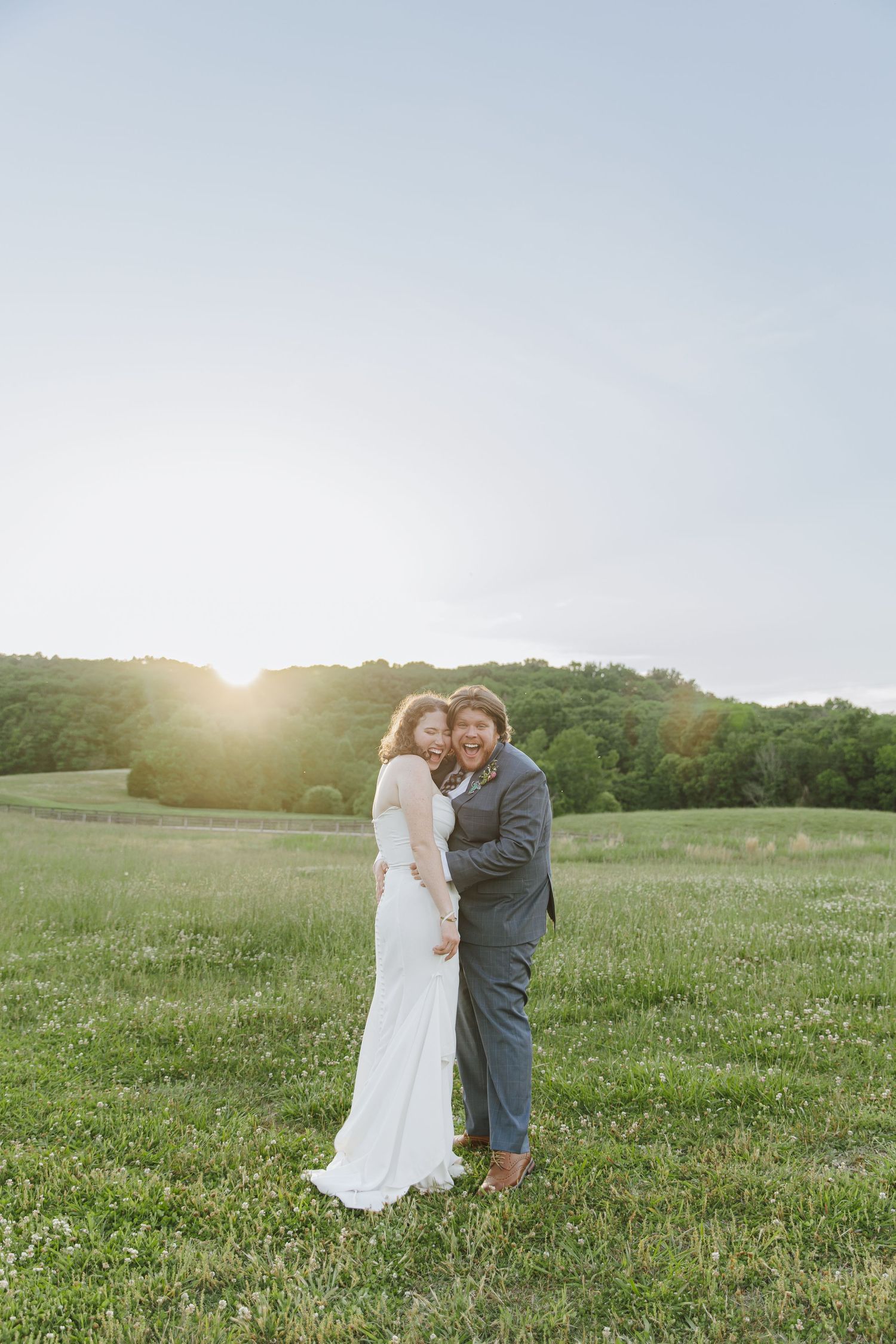 A bride and groom embrace in a sunlit green field at golden hour, with lush trees in the background.