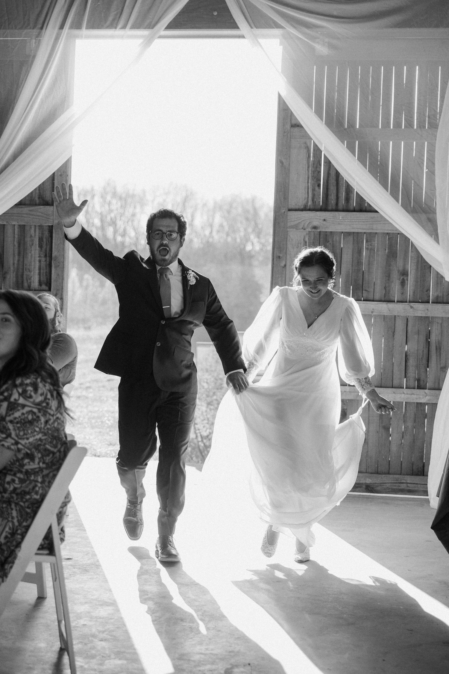 Black and white photo of bride and groom making joyful entrance into rustic barn wedding venue.