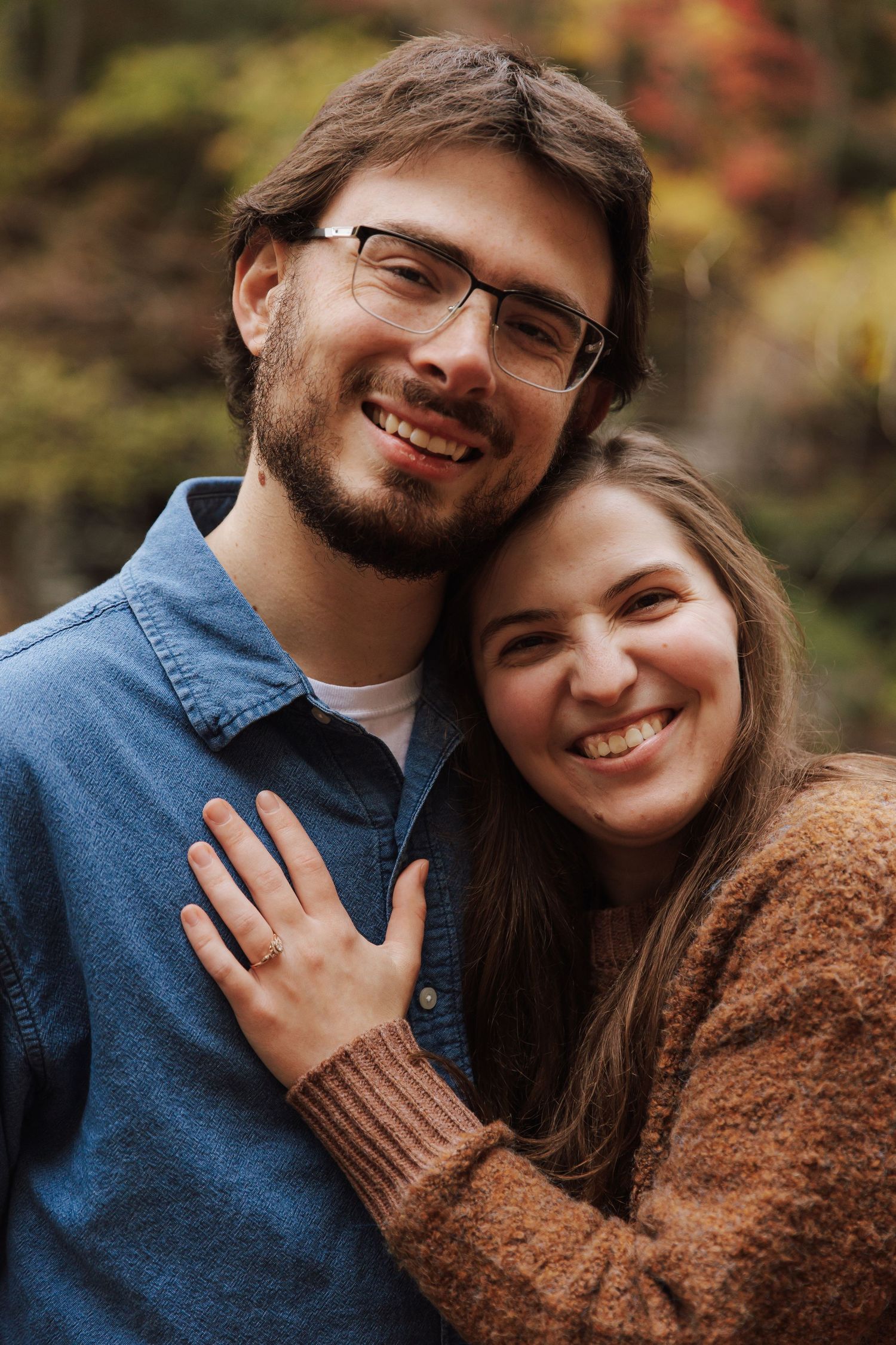 A couple in casual autumn attire embraces and smiles warmly outdoors with fall foliage in the background.