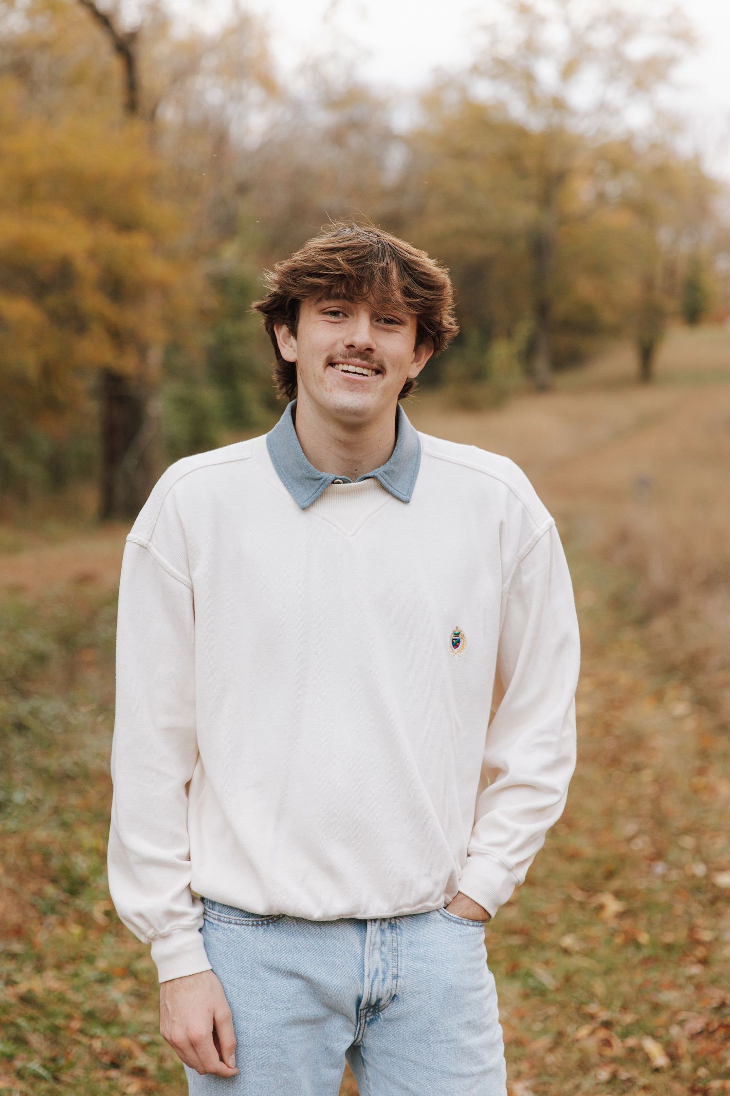 Young person in white sweater and light jeans poses outdoors during autumn season with fallen leaves in background.