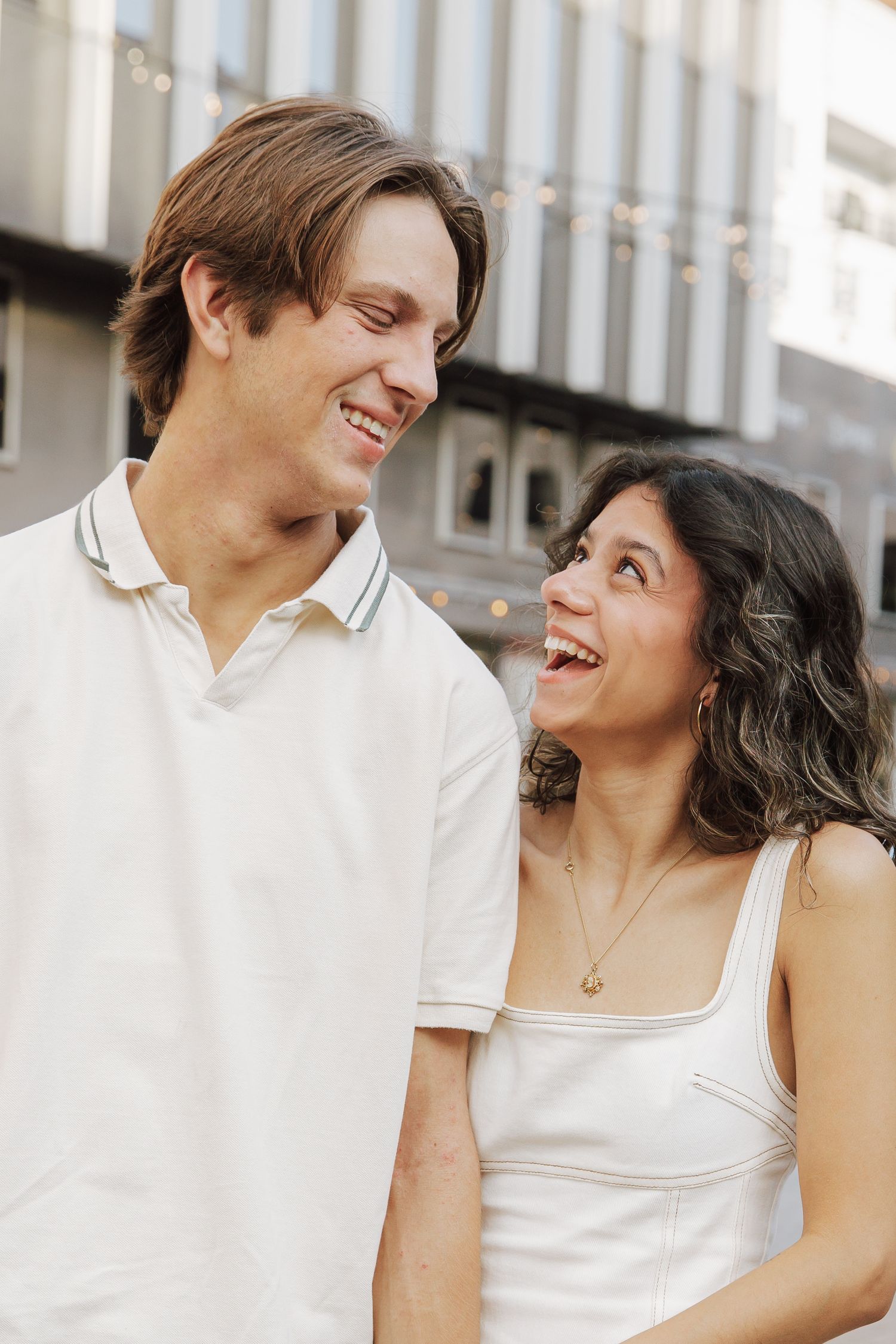 A smiling couple in white outfits laugh together outdoors, with string lights and a building in the background.