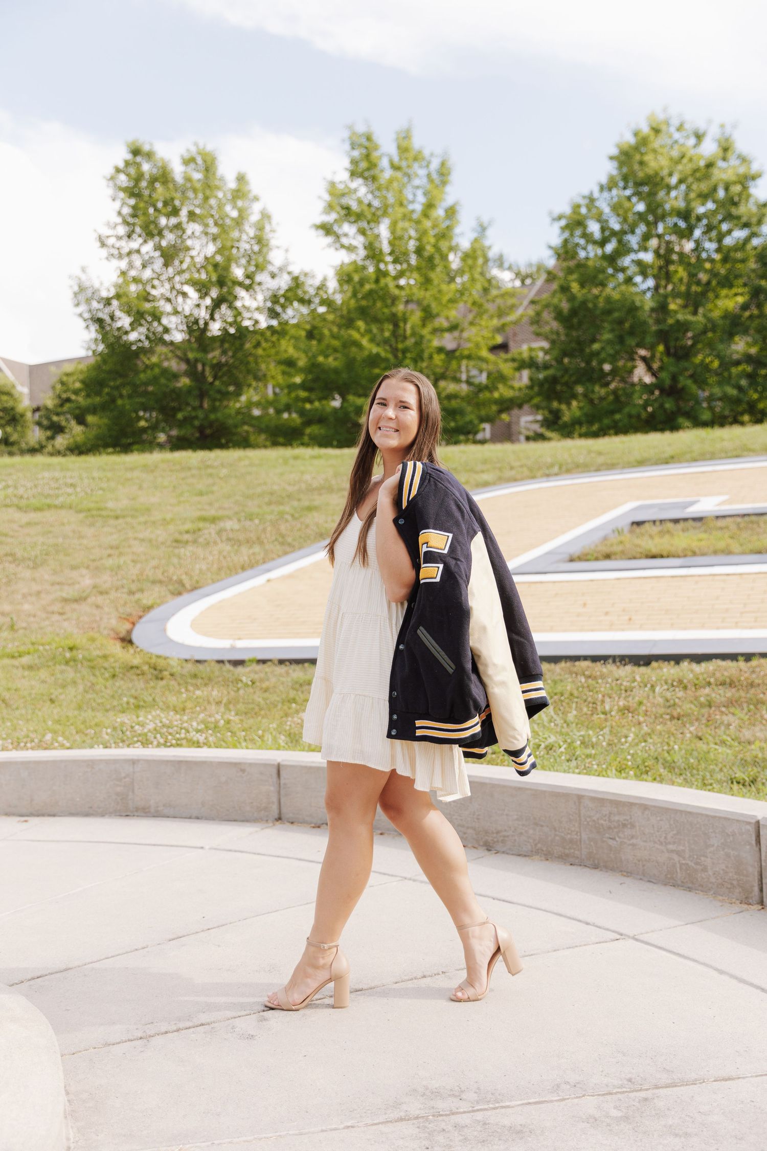 Person in varsity jacket and white dress poses on concrete steps in outdoor setting.