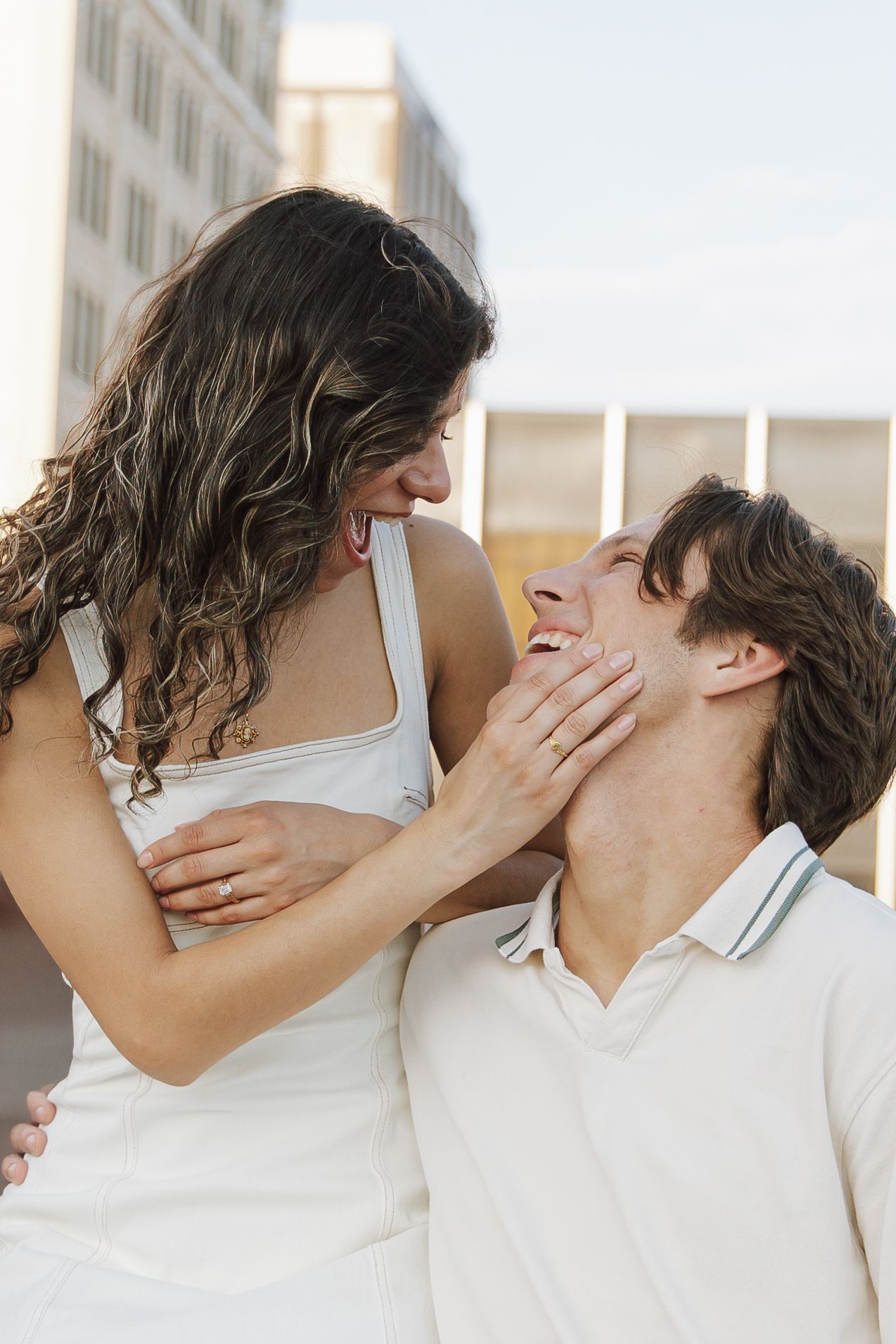 A happy couple in white outfits share a romantic moment outdoors, with the woman tenderly holding the man's face.